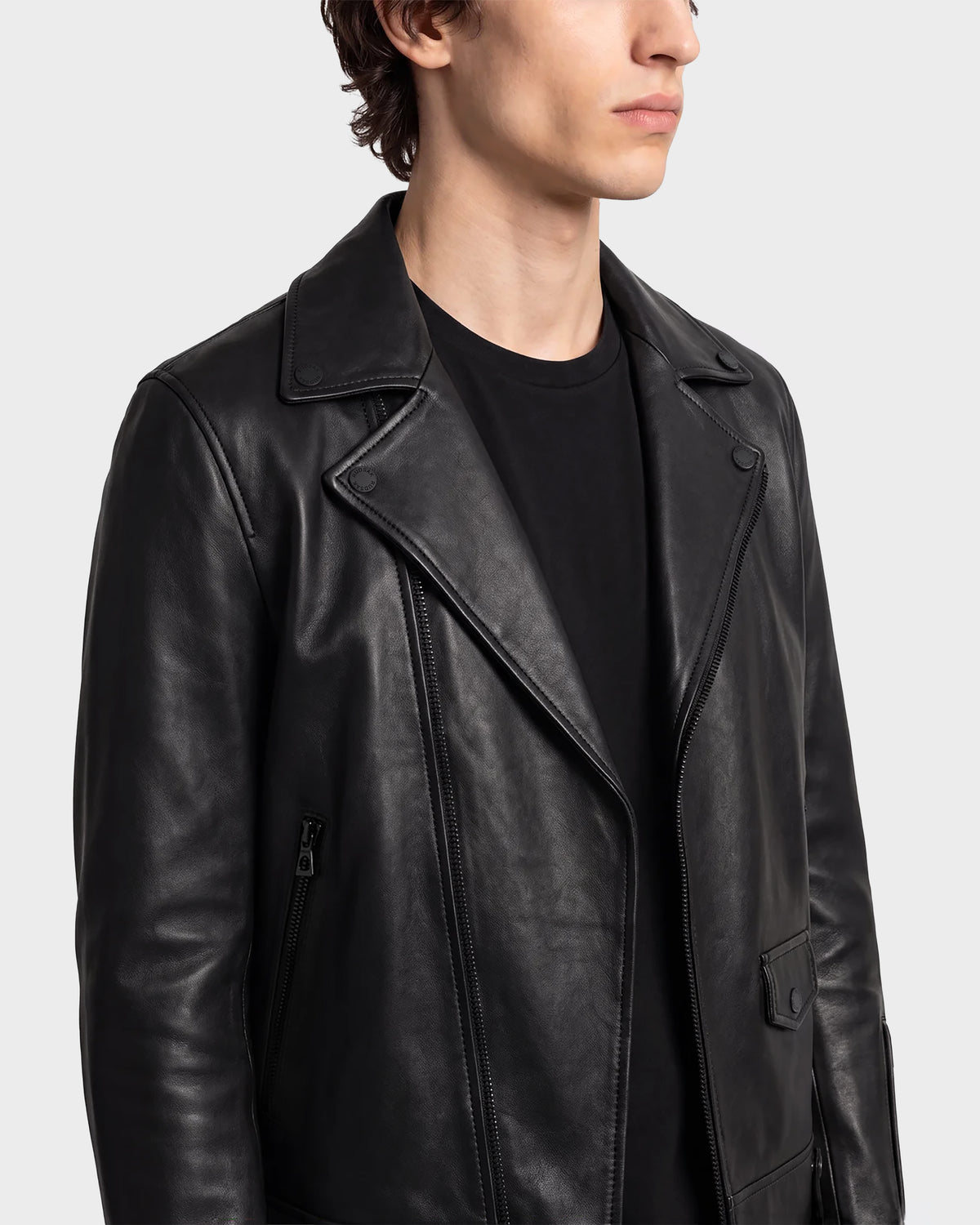 A person wearing the RUDSAK DAREG - 6122004 BLACK biker jacket, expertly crafted from supple lamb leather and layered over a black shirt, faces slightly to the side in a chest-up view against a plain light background. This signature piece embodies effortless style and timeless sophistication.