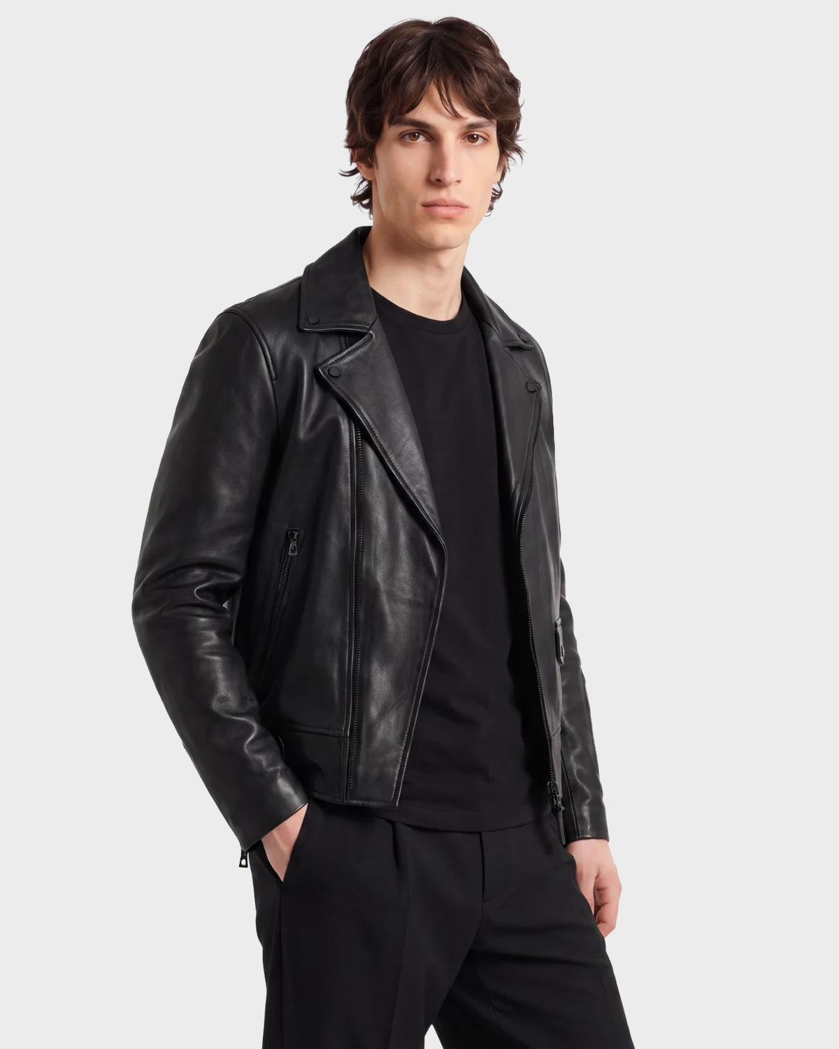 A person with medium-length brown hair wears the RUDSAK DAREG - 6122004 BLACK, a classic black biker jacket crafted from supple lamb leather, layered over a black shirt and black pants and standing against a plain light gray background. This wardrobe essential adds an effortlessly cool edge to the look.