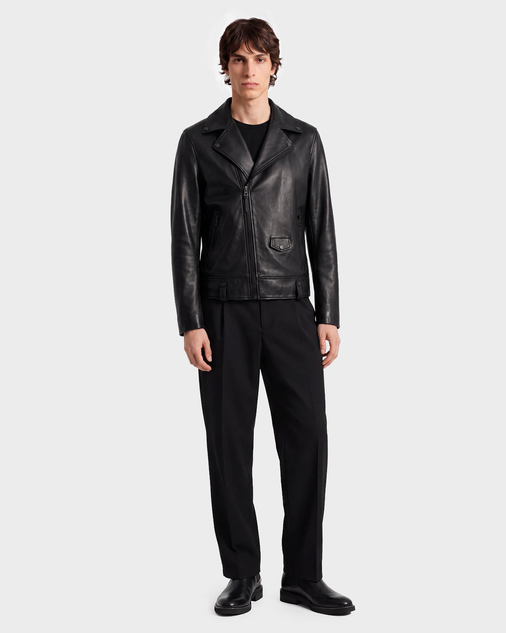 A person stands facing forward against a plain light gray background, wearing the RUDSAK DAREG - 6122004 BLACK lamb leather biker jacket—styled with black pants, black shoes, and a black shirt—showcasing a true wardrobe essential.
