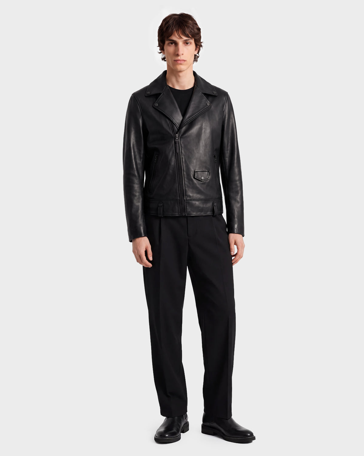 A person stands facing forward against a plain light gray background, wearing the RUDSAK DAREG - 6122004 BLACK lamb leather biker jacket—styled with black pants, black shoes, and a black shirt—showcasing a true wardrobe essential.