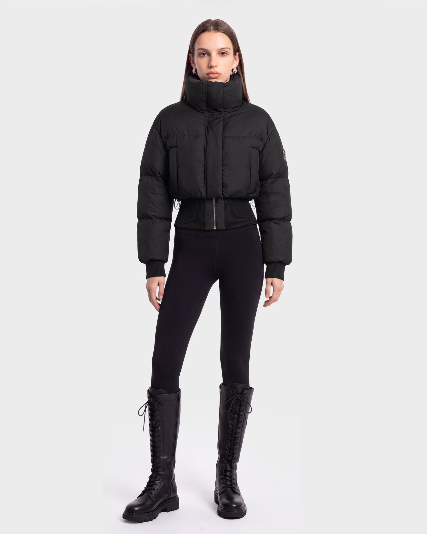 A person stands facing forward, wearing the Rudsak DONYA - 8126516 Black puffer jacket, styled with black leggings and tall black lace-up boots against a plain light gray background. This look is available for pre-order and is expected to ship by September 15th.