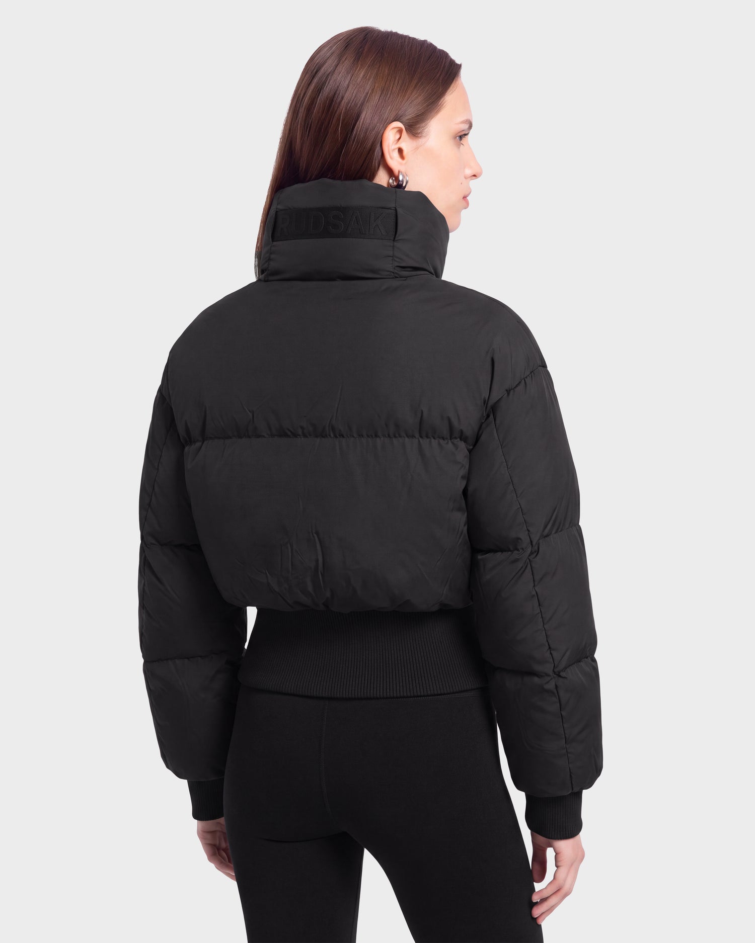 A woman with straight brown hair models the Rudsak DONYA - 8126516 Black puffer jacket, featuring a high collar and paired with fitted black pants. She stands with her back to the camera against a plain light background. This stylish look is available for pre-order and is expected to ship by September 15th.