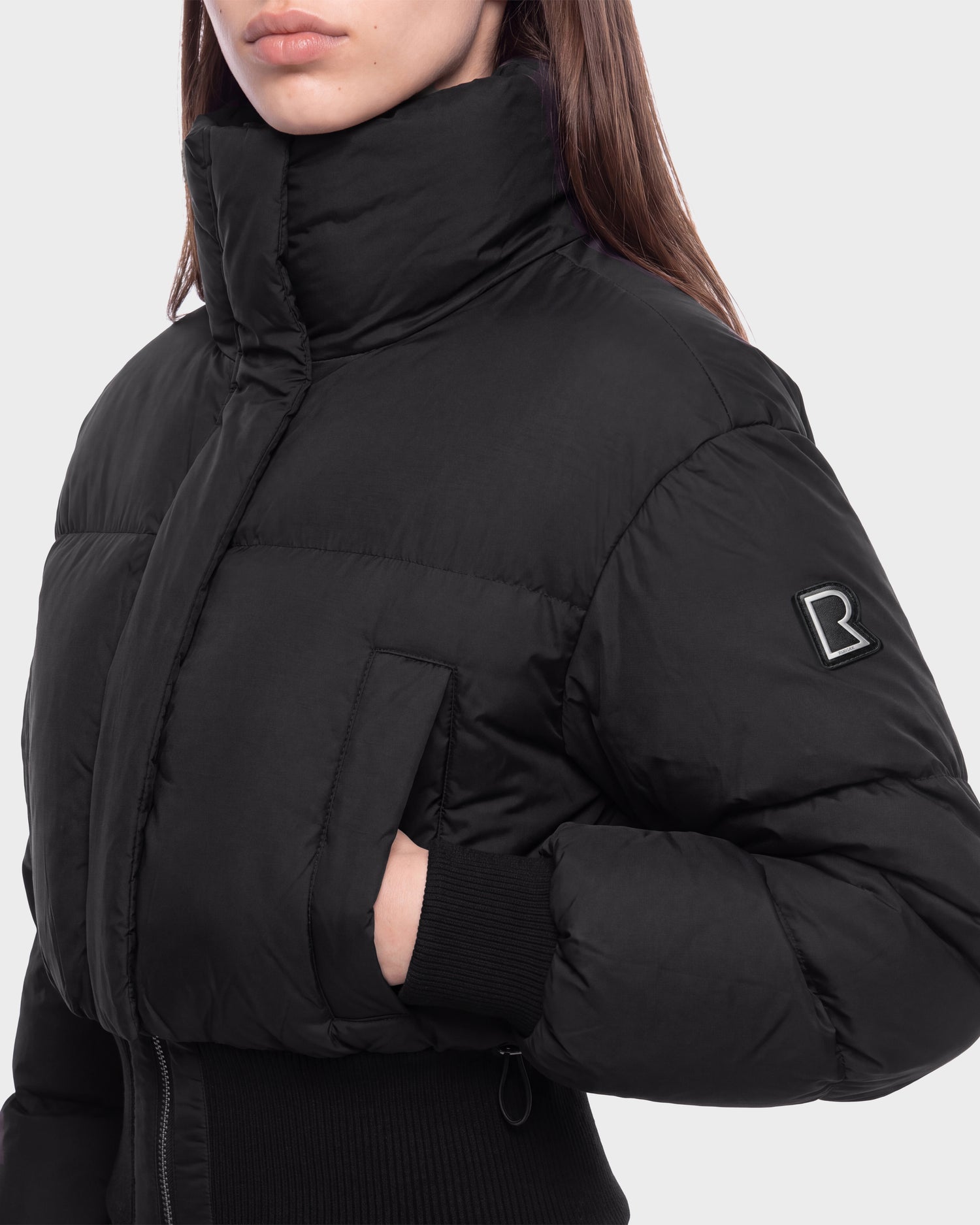 A woman models the Rudsak DONYA - 8126516 Black puffer jacket, which features a high collar, side pocket, and a distinctive B logo patch on the sleeve. Only her lower face and upper body are shown. This jacket is available for pre-order and is expected to ship by September 15th.