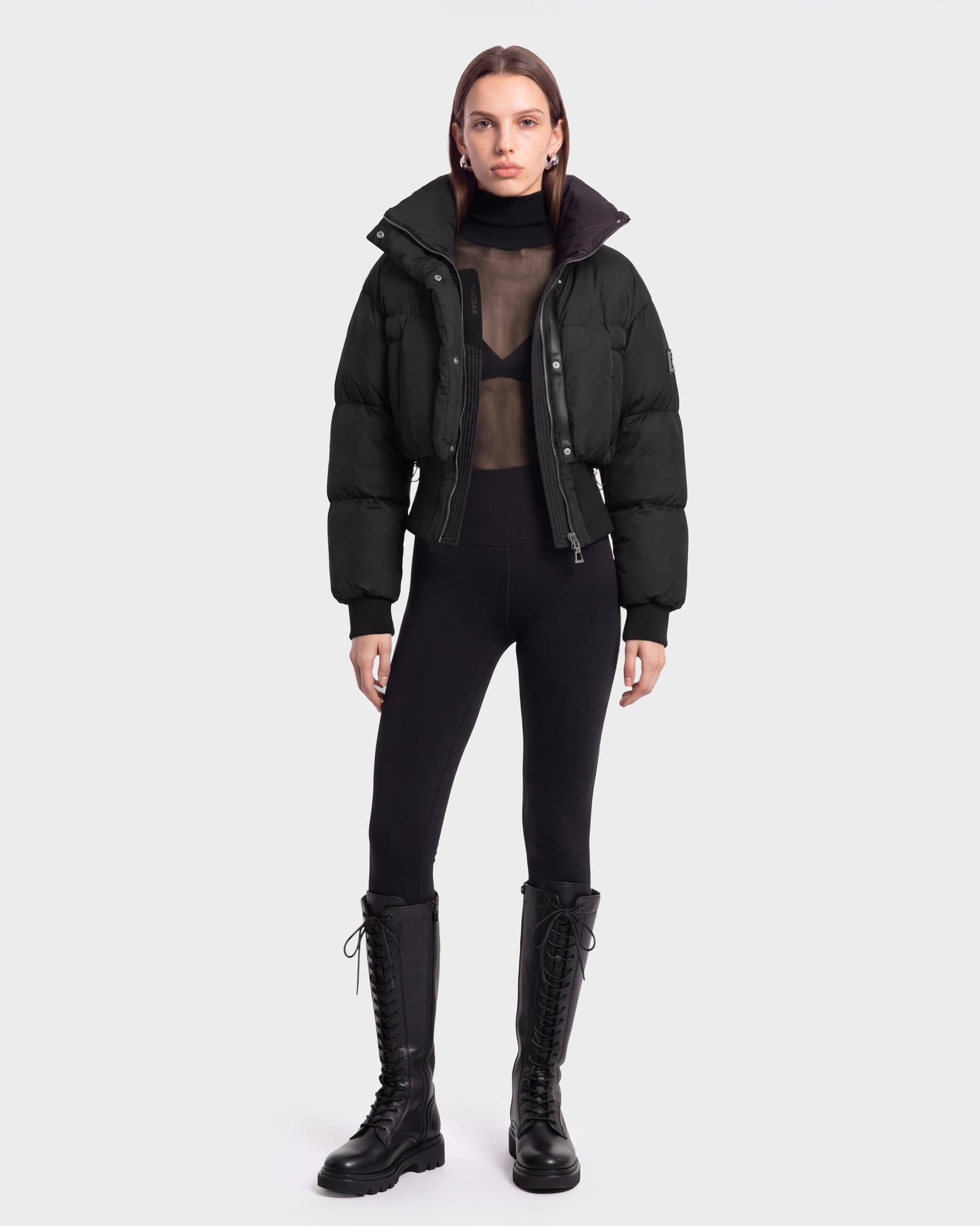 A model stands facing forward against a plain white background, wearing the Rudsak DONYA - 8126516 Black puffer jacket styled with a sheer black top, black leggings, and tall lace-up boots—a look now available for pre-order and expected to ship by September 15th.
