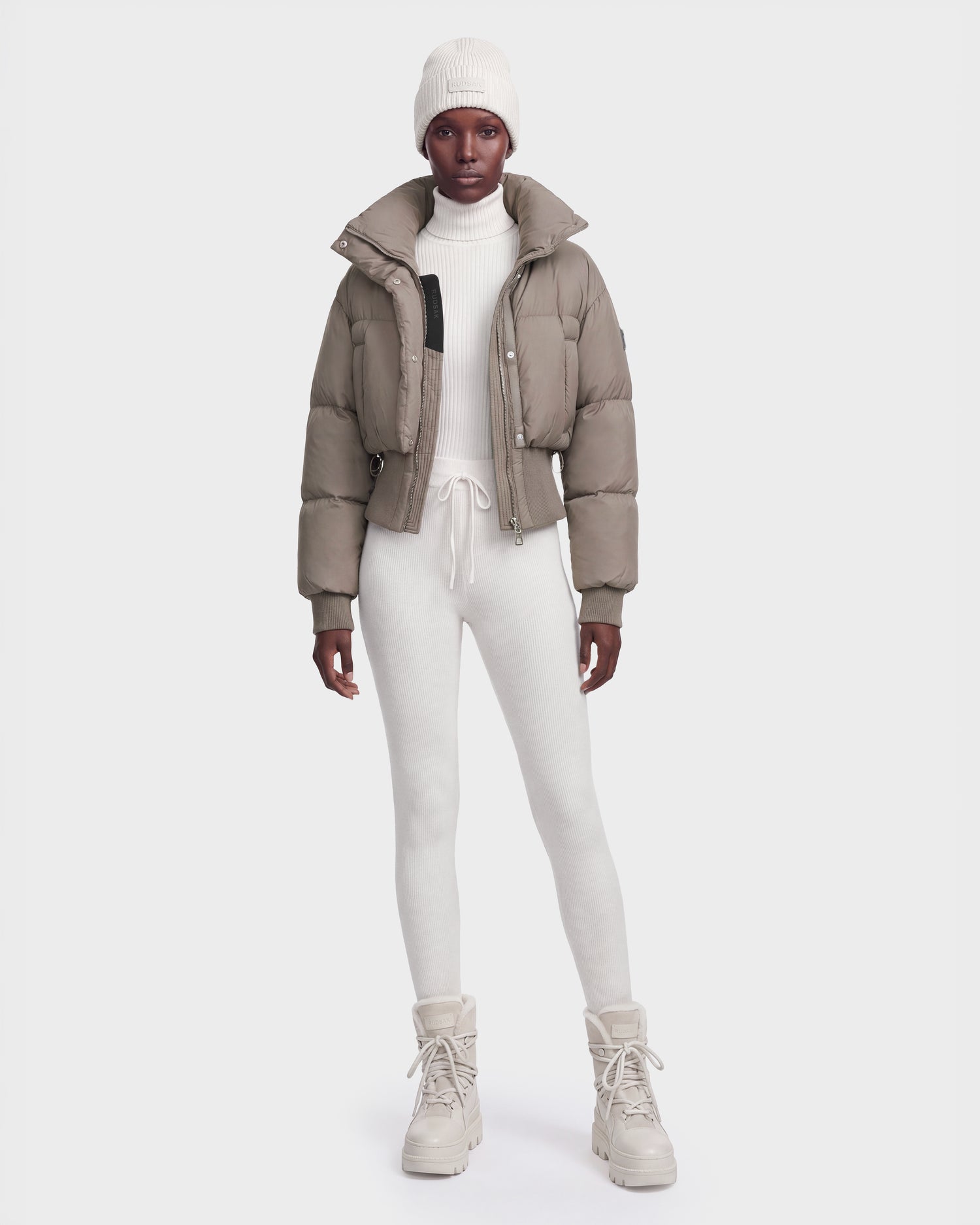 A person wearing the Rudsak DONYA - 8126516 Cement women’s down puffer jacket filled with RDS-certified down, paired with a white knit beanie, a white turtleneck, white leggings, and beige lace-up boots stands against a plain white background.