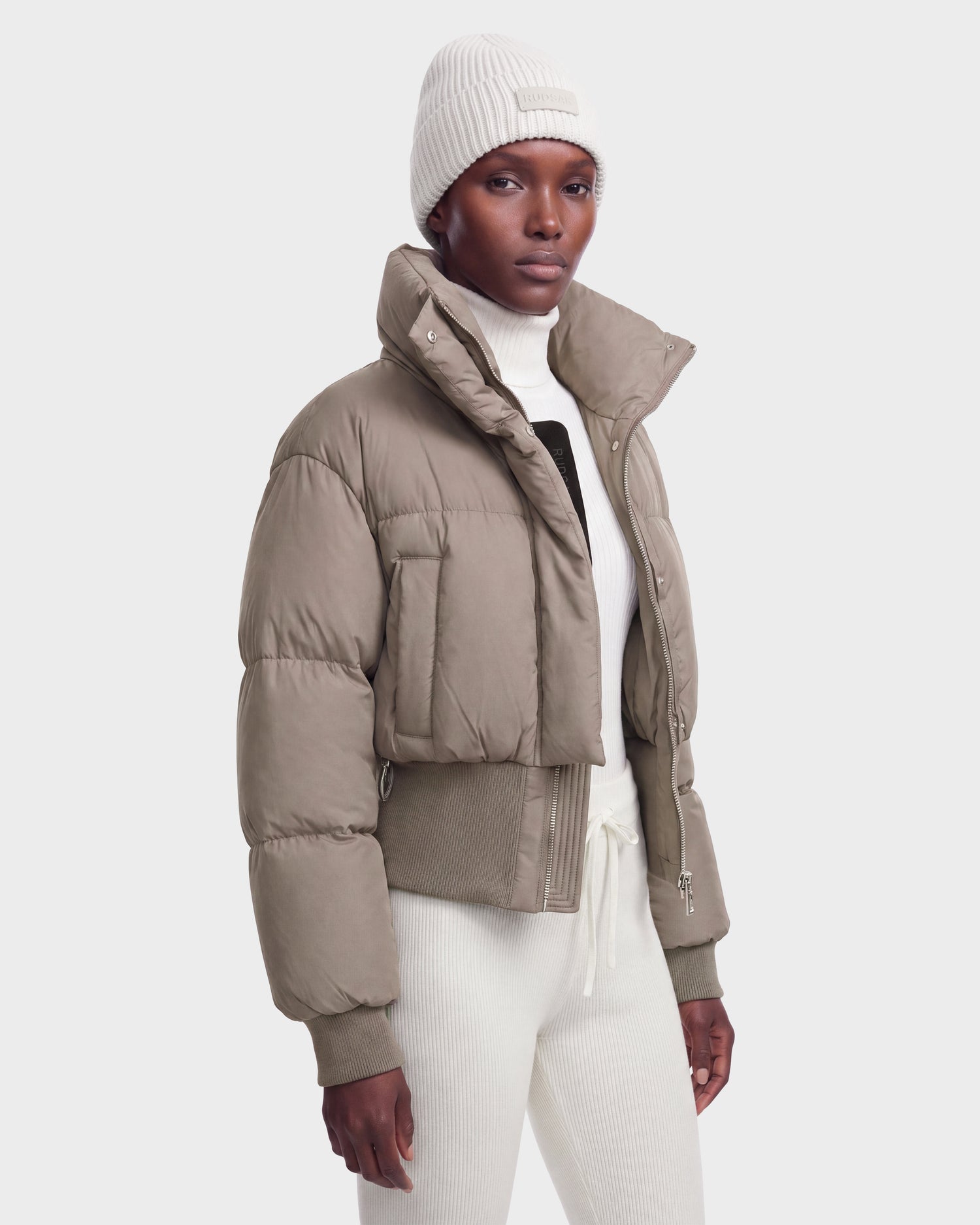 A person wearing the Rudsak DONYA - 8126516 Cement women’s down puffer jacket, filled with RDS-certified down, along with a white knit beanie, white ribbed turtleneck sweater, and white pants stands against a plain light background.
