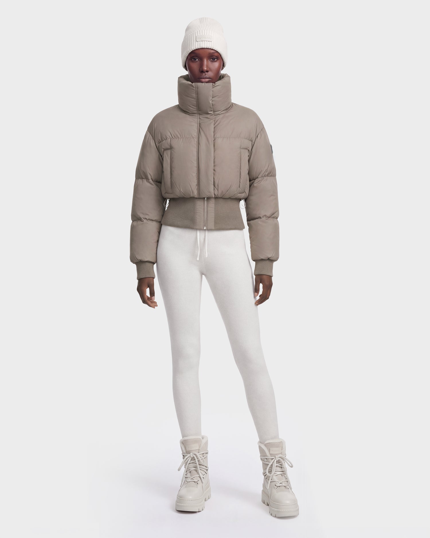 A person wearing the Rudsak DONYA - 8126516 Cement women’s down puffer jacket, filled with RDS-certified down, white leggings, beige lace-up boots, and a white knit beanie stands facing forward against a plain white background.