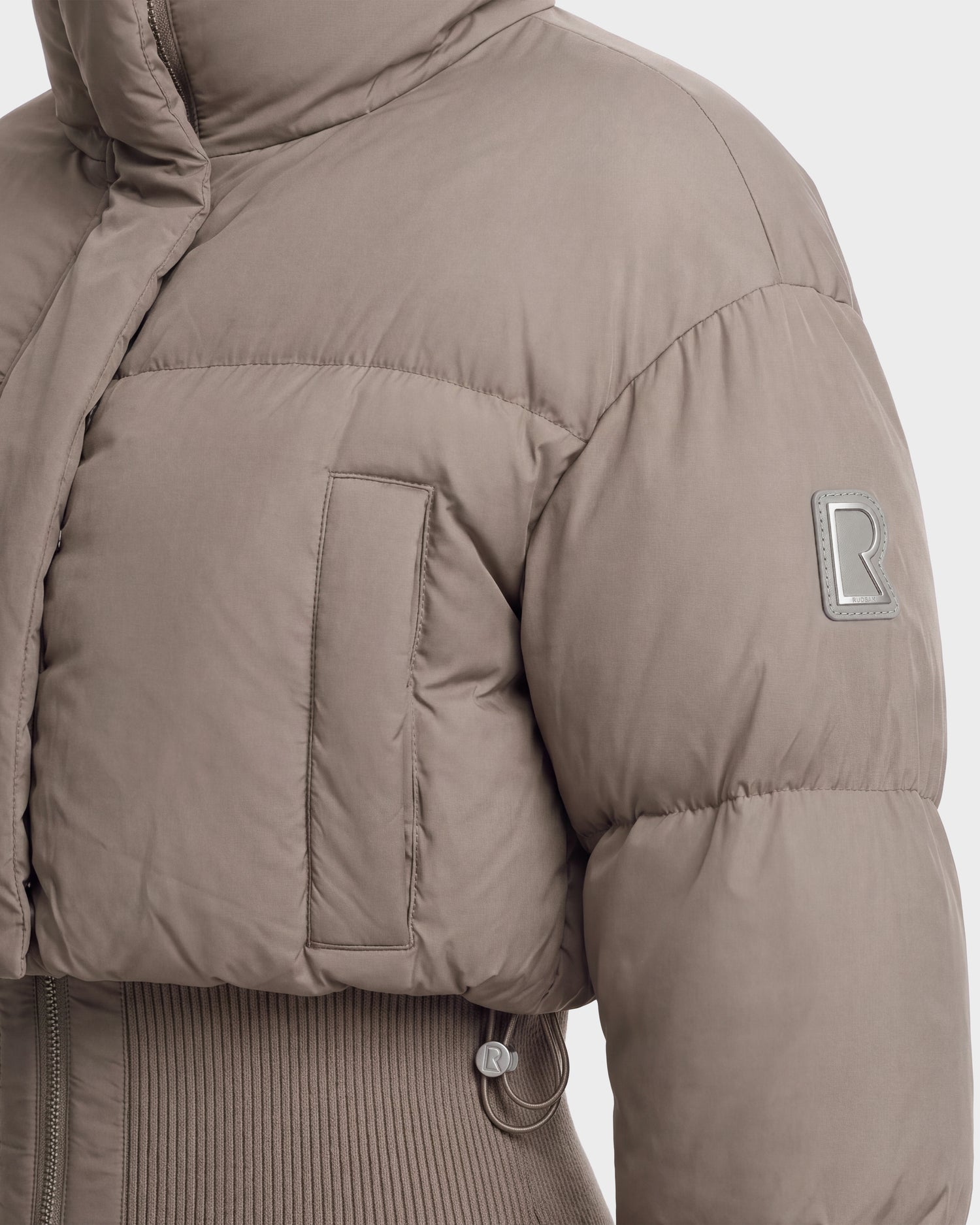 Close-up of the DONYA - 8126516 Cement women’s down puffer jacket by Rudsak in taupe. This winter jacket features a ribbed hem, chest pocket, and an R logo patch on the sleeve. Crafted from matte, soft-textured fabric and filled with RDS-certified down for added warmth, it also offers a high collar for extra protection against the cold.