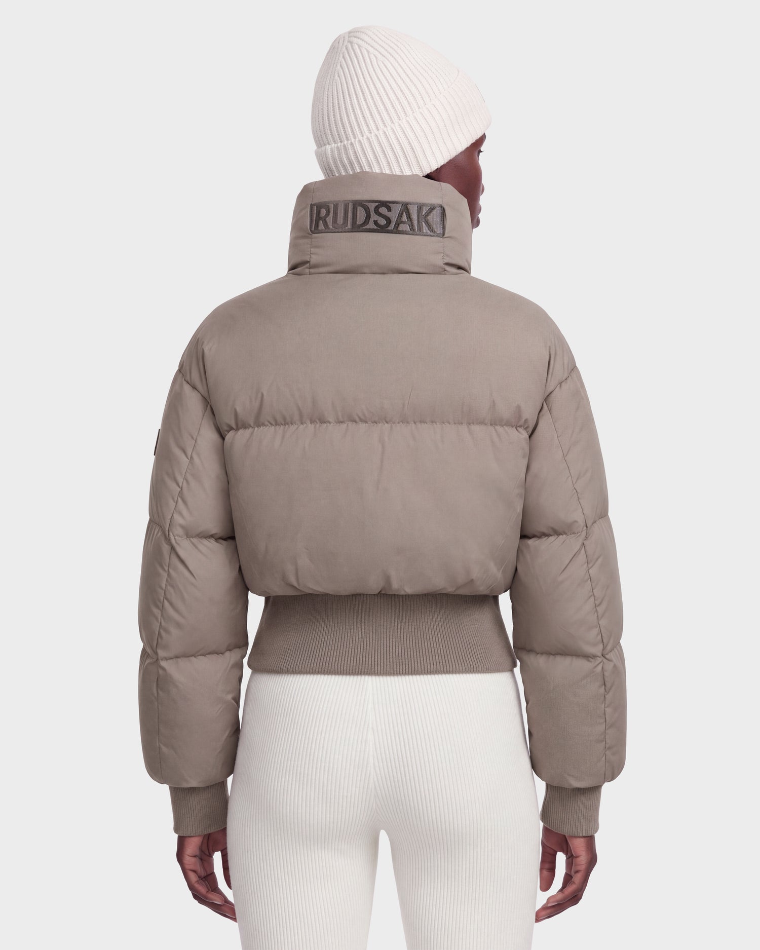 A woman is shown from the back against a plain light background, wearing Rudsak’s DONYA - 8126516 Cement women’s down puffer jacket featuring RUDSAK branding on the high collar, paired with white ribbed pants and a white knit beanie. This RDS-certified winter jacket combines warmth and style for comfortable cold-weather wear.