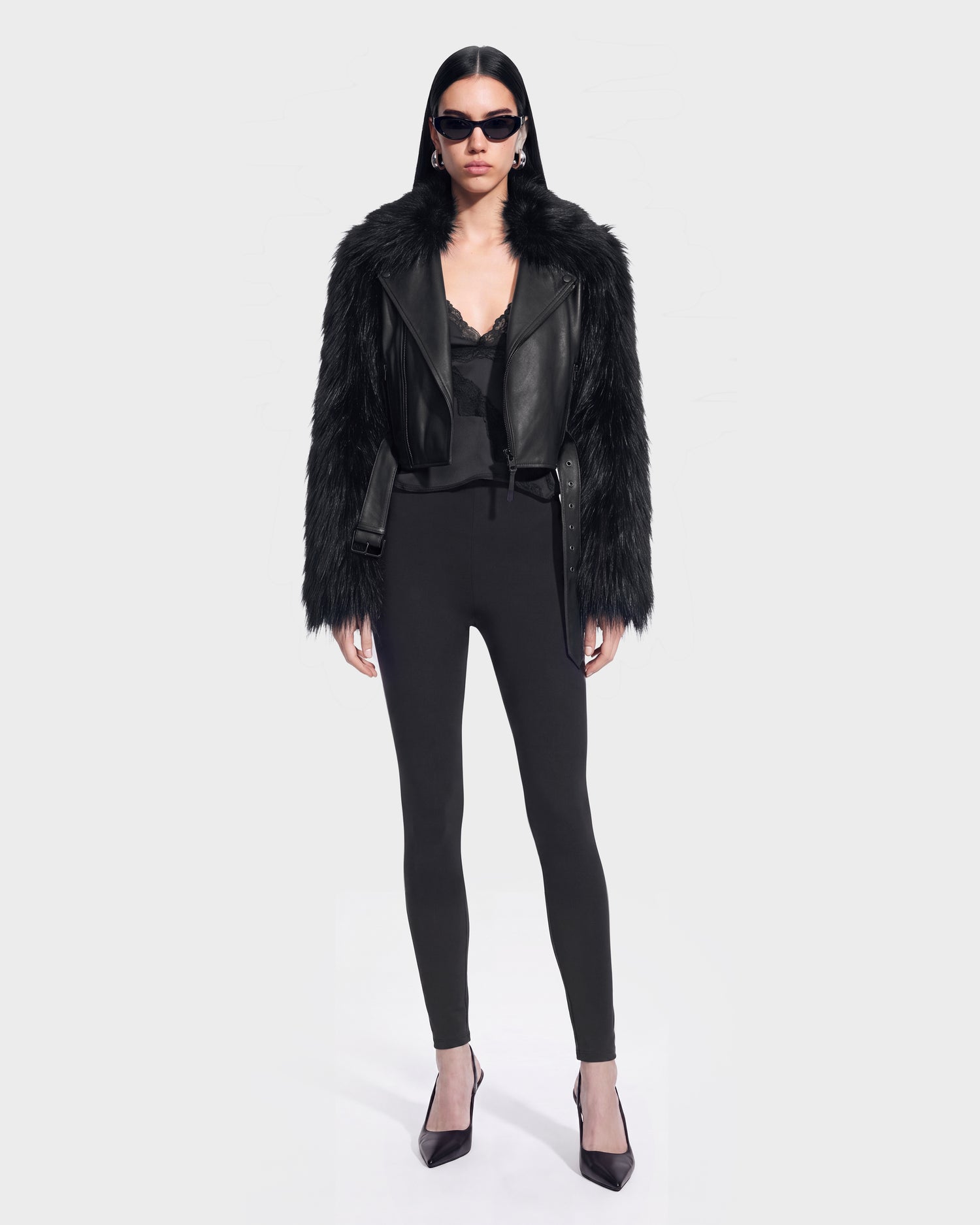 A woman wears the EMELDA - 8126991 Black jacket by Rudsak—a bold black faux fur and leather piece—styled with a black lace top, high-waisted black pants, pointed black heels, and sunglasses against a plain light background. This striking look is available for pre-order now and is expected to ship by September 15th.