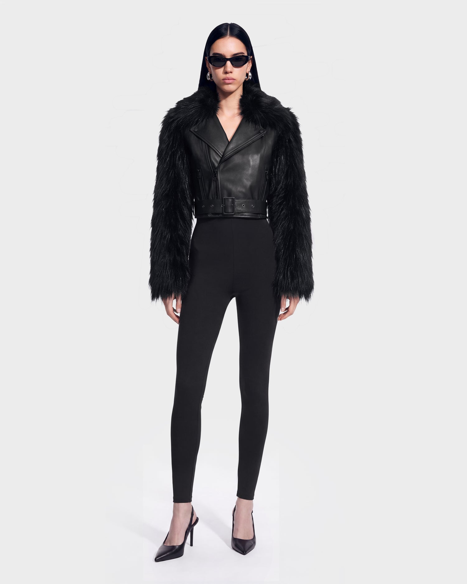 A person dressed in Rudsak’s EMELDA - 8126991 Black—featuring black sunglasses, a belted leather jacket with furry sleeves, fitted black pants, and sleek black heels—stands against a plain, light background; this bold look is available for pre-order now and will ship by September 15th.