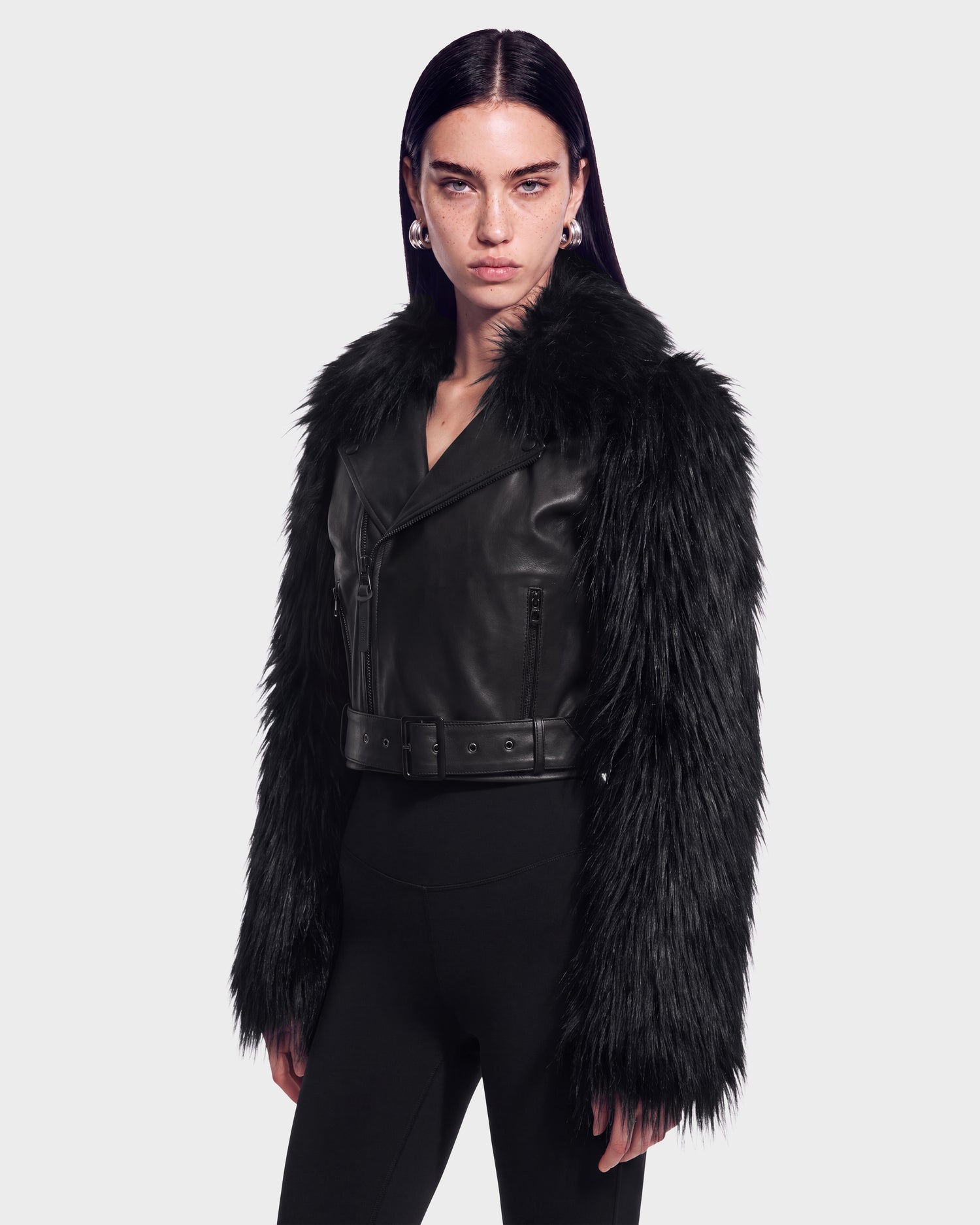 A woman wearing the Rudsak EMELDA - 8126991 Black leather jacket, featuring a faux fur collar and sleeves, paired with black pants and silver hoop earrings, stands against a plain white background. This stylish jacket is available for pre-order now and is expected to ship by September 15th.