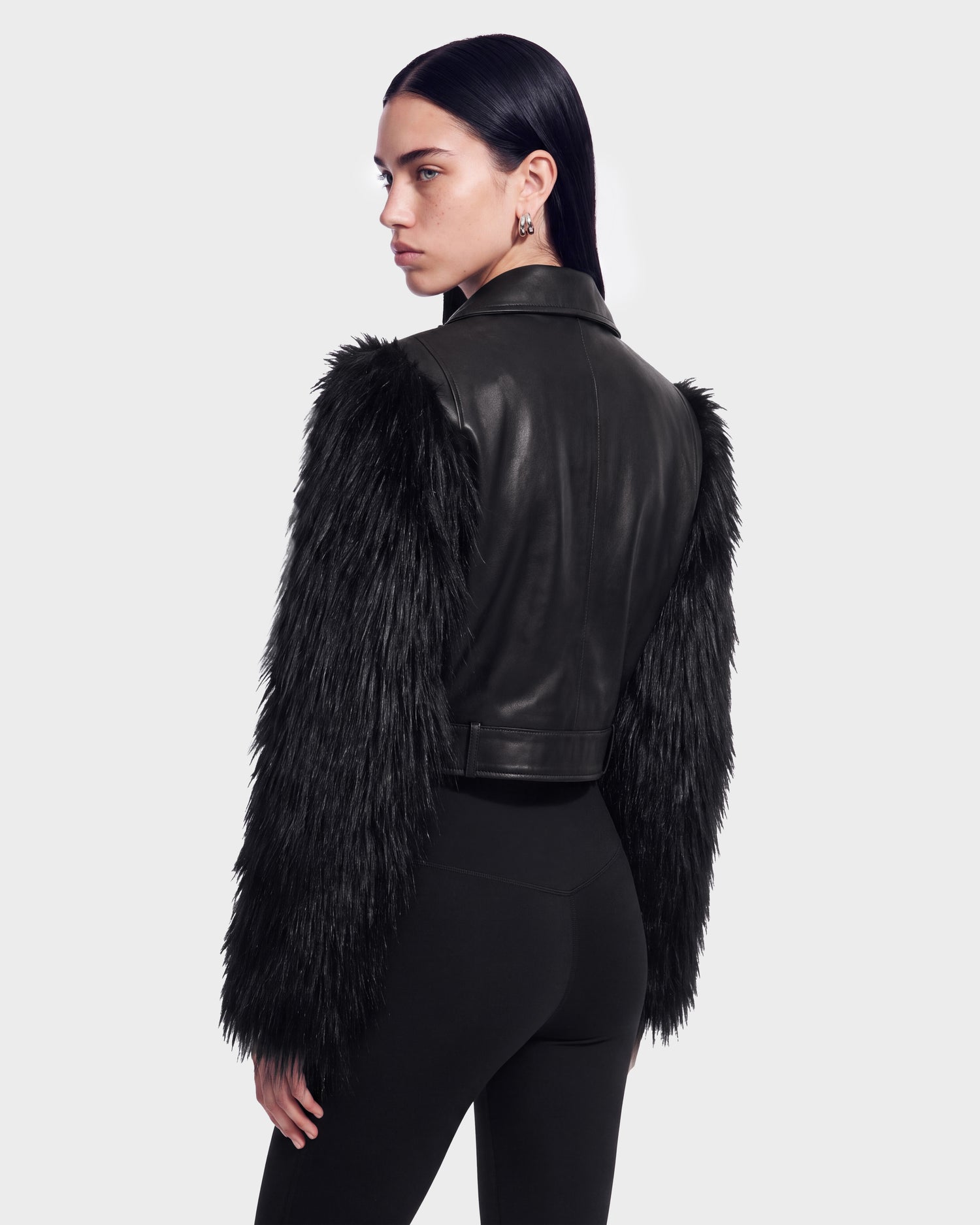 A woman wearing the EMELDA - 8126991 Black leather jacket with furry sleeves from Rudsak and black pants stands looking over her shoulder against a plain light background. This stylish look is available for pre-order and is expected to ship by September 15th.