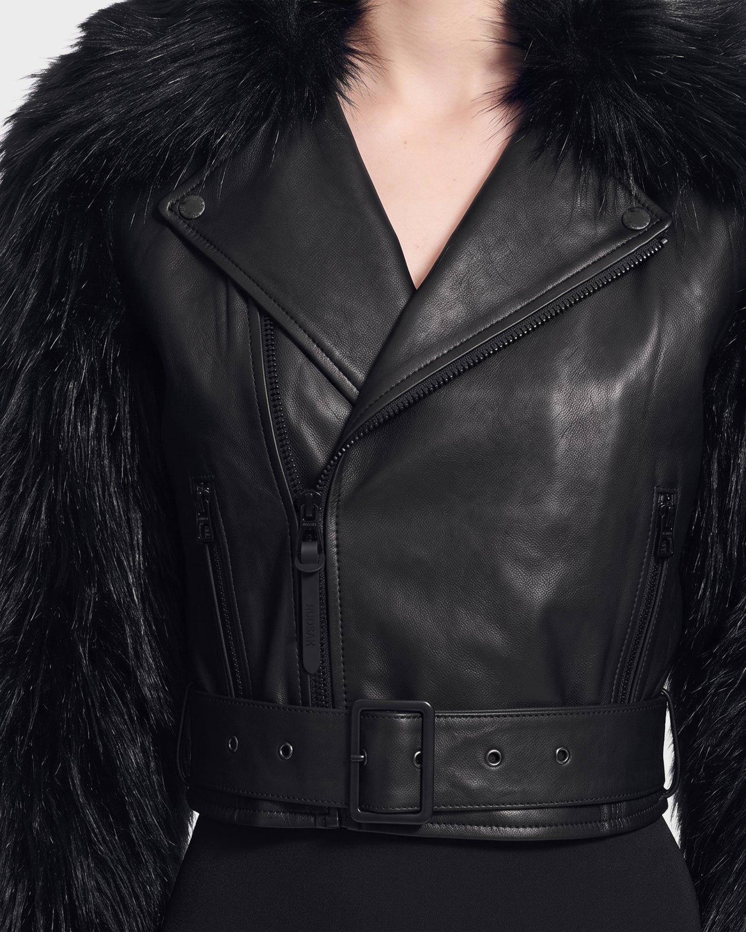 A person is shown from the neck to the waist, wearing the Rudsak EMELDA - 8126991 Black leather jacket, which features a wide belt and faux fur trim on the shoulders, set against a white background. This stylish piece is available for pre-order and is expected to ship by September 15th.