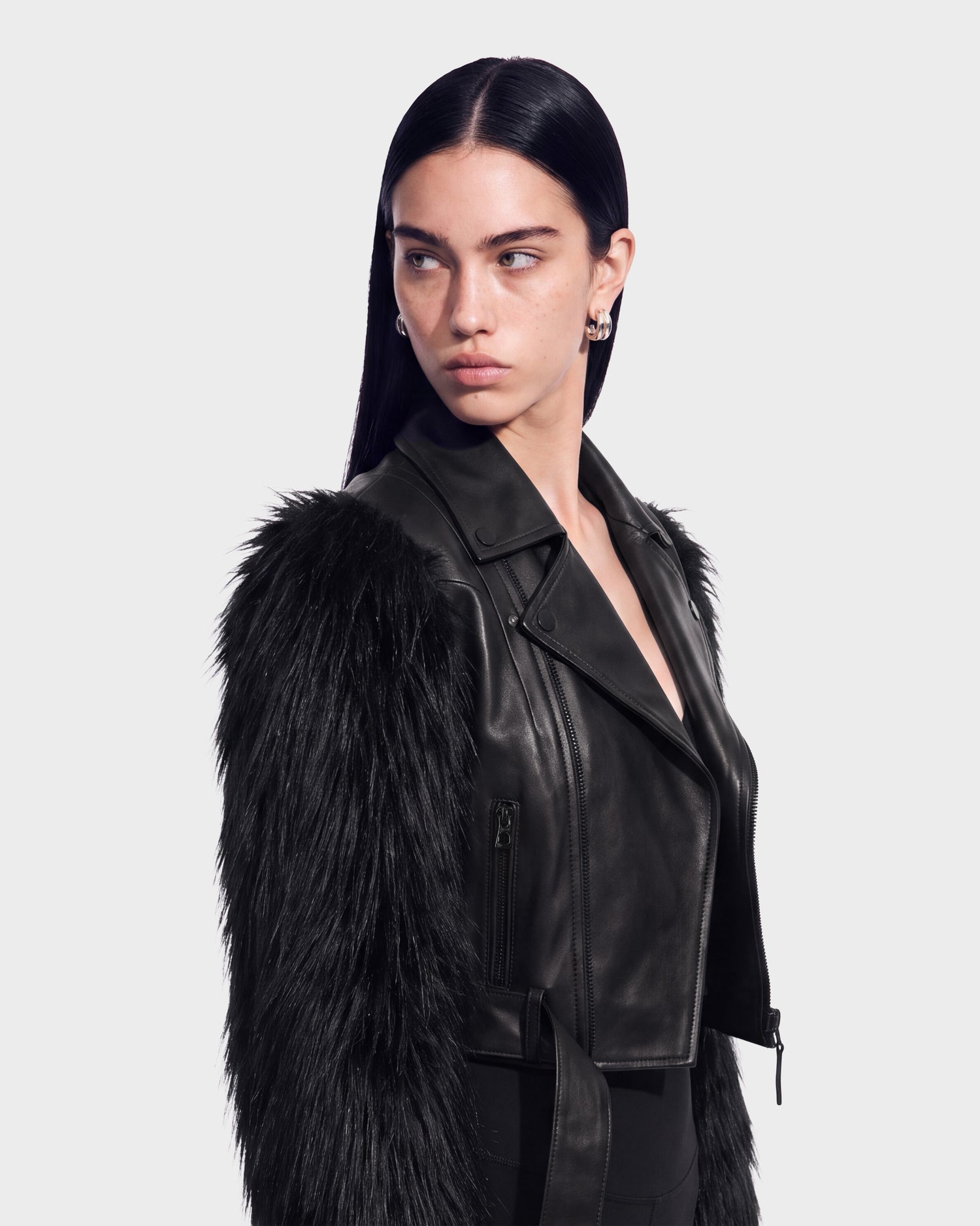 A woman with long dark hair, wearing the Rudsak EMELDA - 8126991 Black faux fur and leather jacket, looks to her left against a plain light background. This chic piece is available for pre-order now and is expected to ship by September 15th.