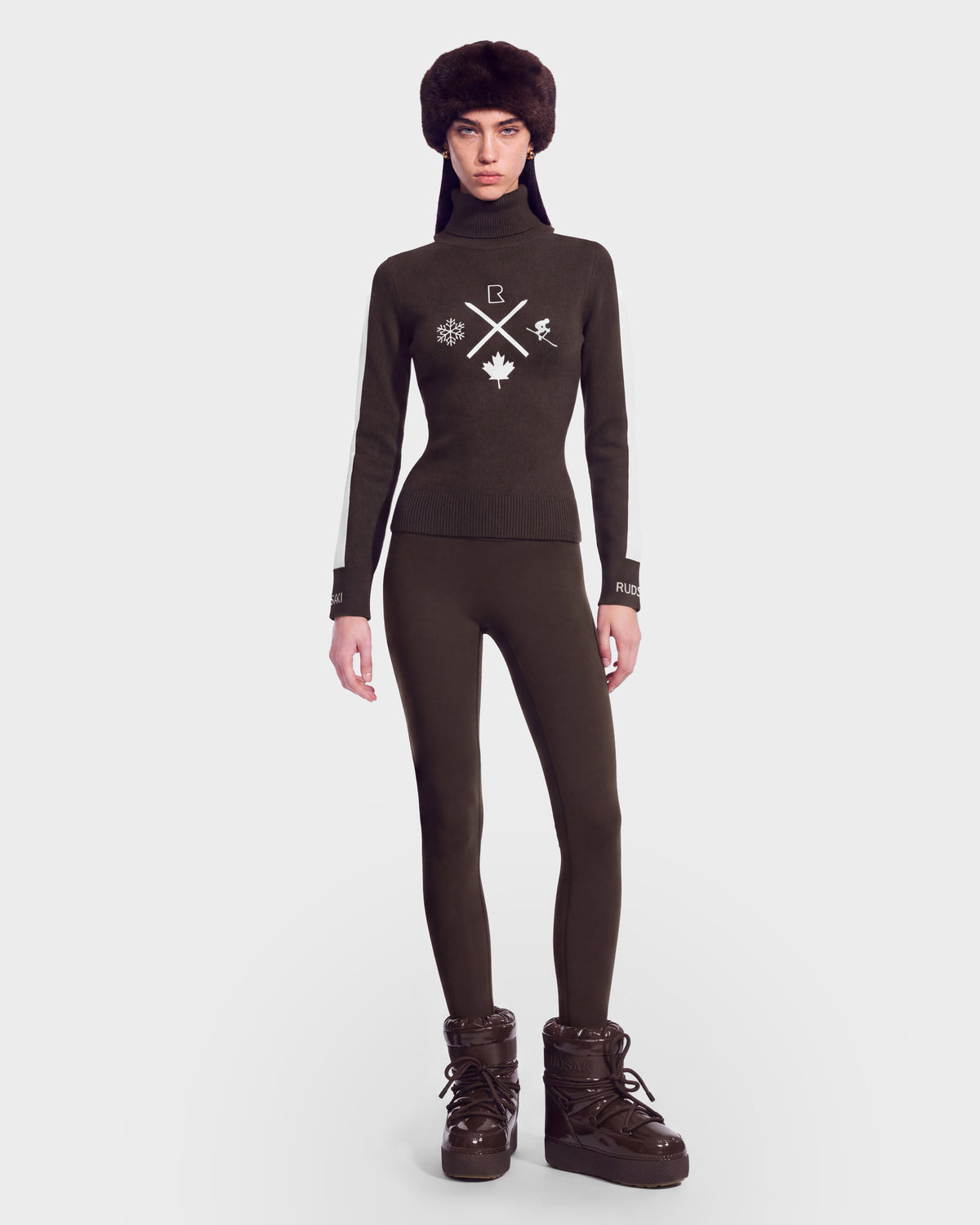 A person wearing a dark fur hat, a Rudsak women’s ski turtleneck with winter-themed symbols, fitted black pants, and the ELIZABETH - 8524623 Espresso brown winter boots by Rudsak stands against a plain white background—an effortlessly stylish look for your winter wardrobe.