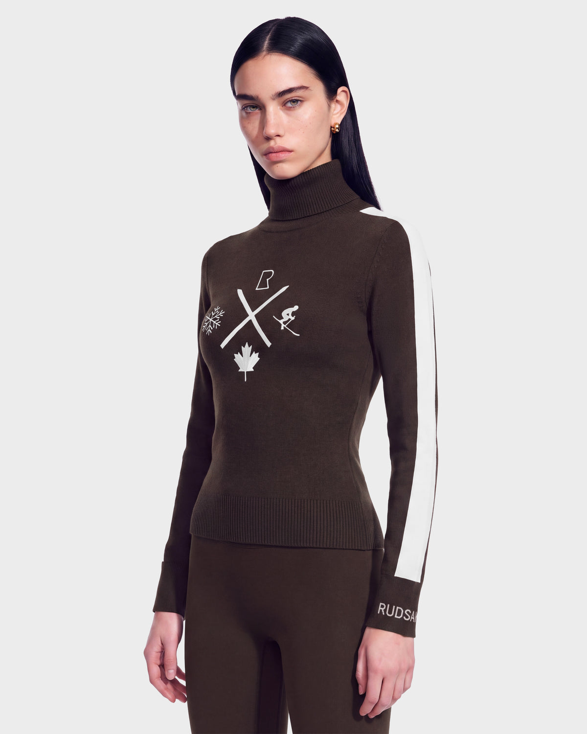 A woman with long dark hair wears the Rudsak ELIZABETH - 8524623 Espresso women’s ski turtleneck sweater, featuring white symbols and a white stripe down the sleeve, paired with matching brown pants, standing against a plain background—an effortless addition to any winter wardrobe.