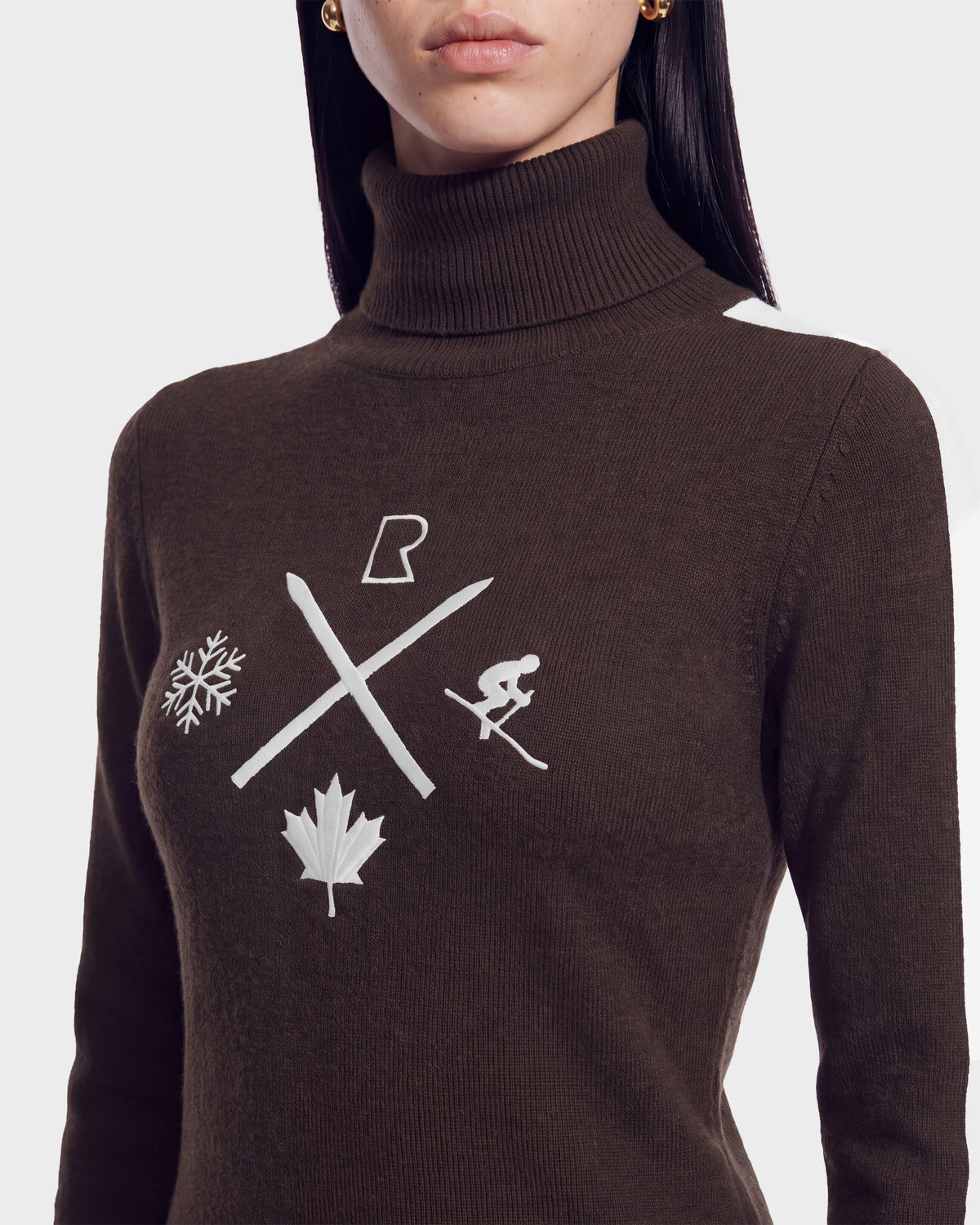 A woman wearing the ELIZABETH - 8524623 Espresso women’s ski turtleneck from Rudsak, featuring a rich brown color and distinctive white symbols on the front—including a snowflake, crossed skis, a skier, a maple leaf, and a stylized letter R—making it an ideal choice for your winter wardrobe or après-ski gatherings.