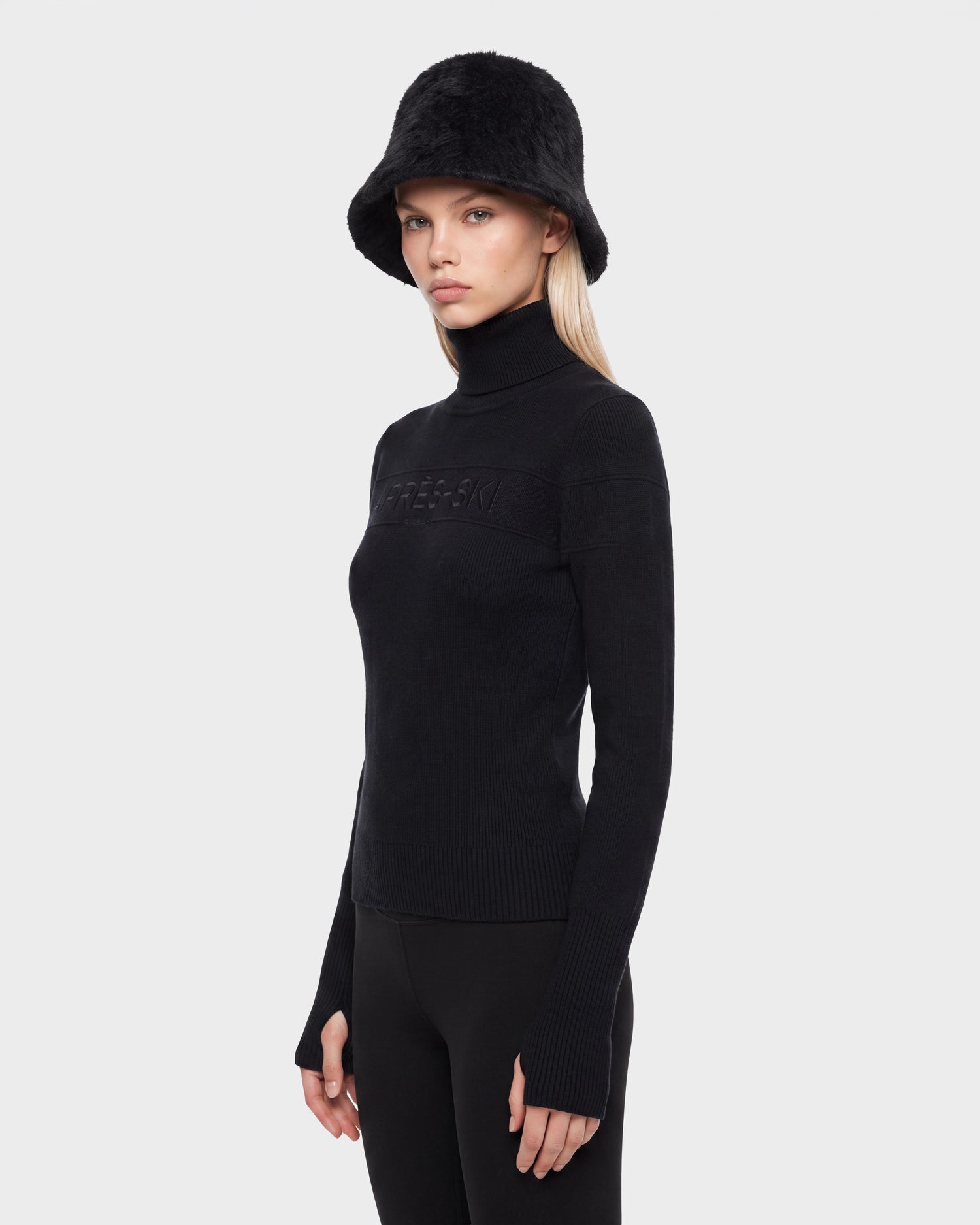 A woman stands against a plain light grey background, wearing the Rudsak ERIKA - 8525705 Black turtleneck sweater paired with black pants and a black fuzzy bucket hat. Looking slightly to the side with a neutral expression, she effortlessly highlights this essential winter piece from Rudsak.