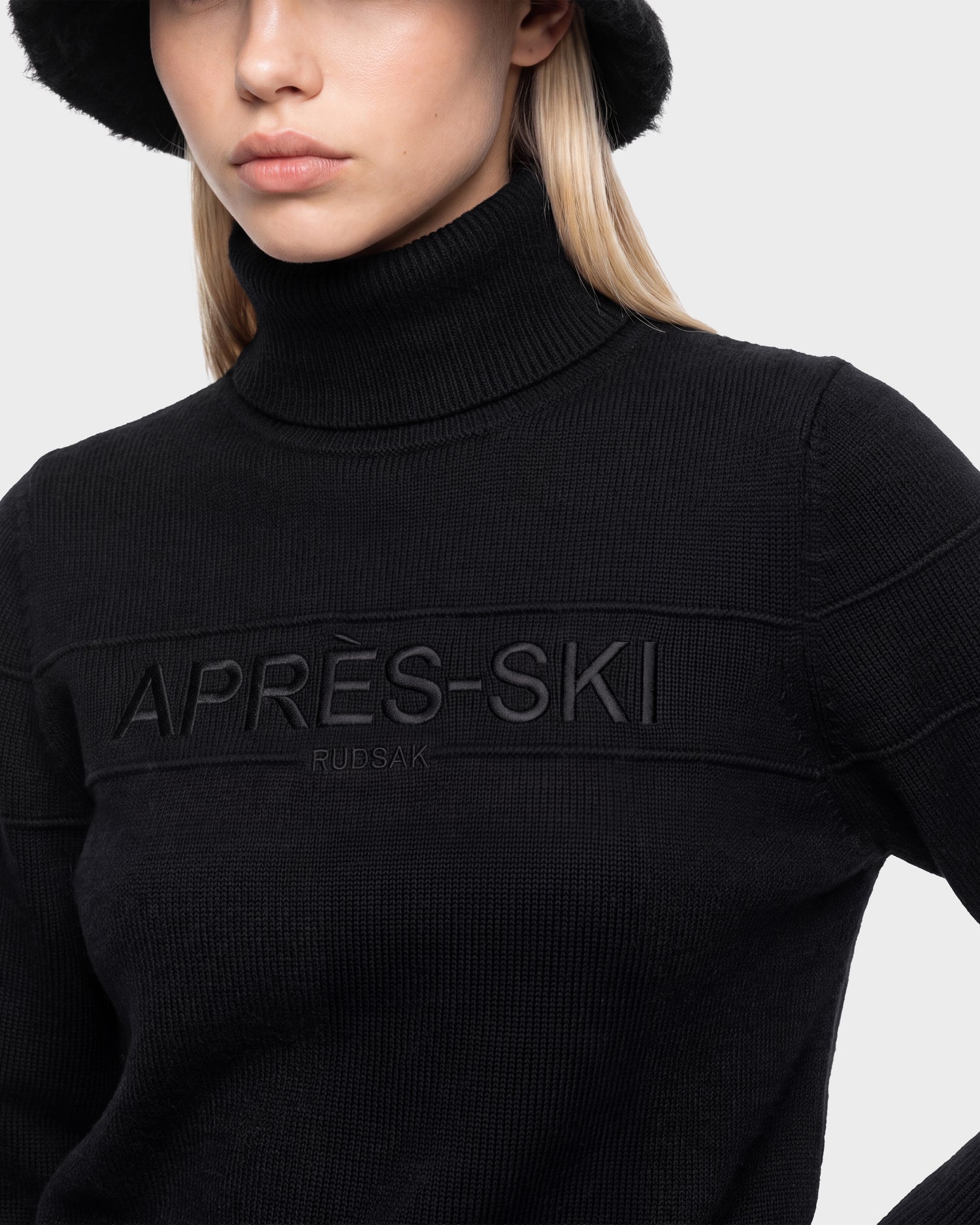 Person wearing the ERIKA - 8525705 Black sweater by Rudsak, featuring an embossed APRÈS-SKI RUBSAK detail on the chest for a classic après-ski vibe, paired with a black bucket hat and posing against a plain light background.