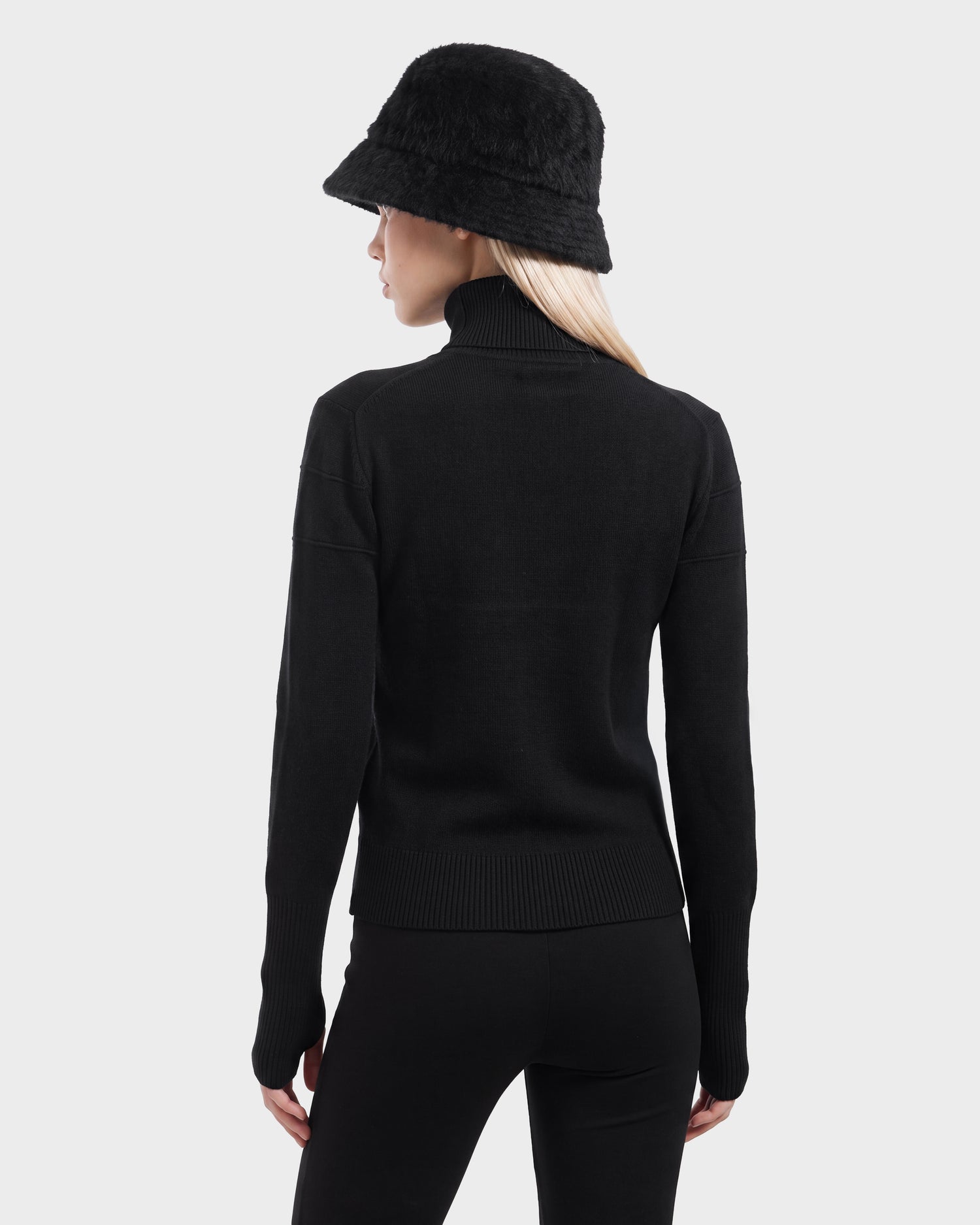 A woman stands facing away, dressed in the Rudsak ERIKA - 8525705 Black turtleneck sweater—a timeless staple for any winter wardrobe—paired with black pants and a black fuzzy bucket hat against a plain light gray background.
