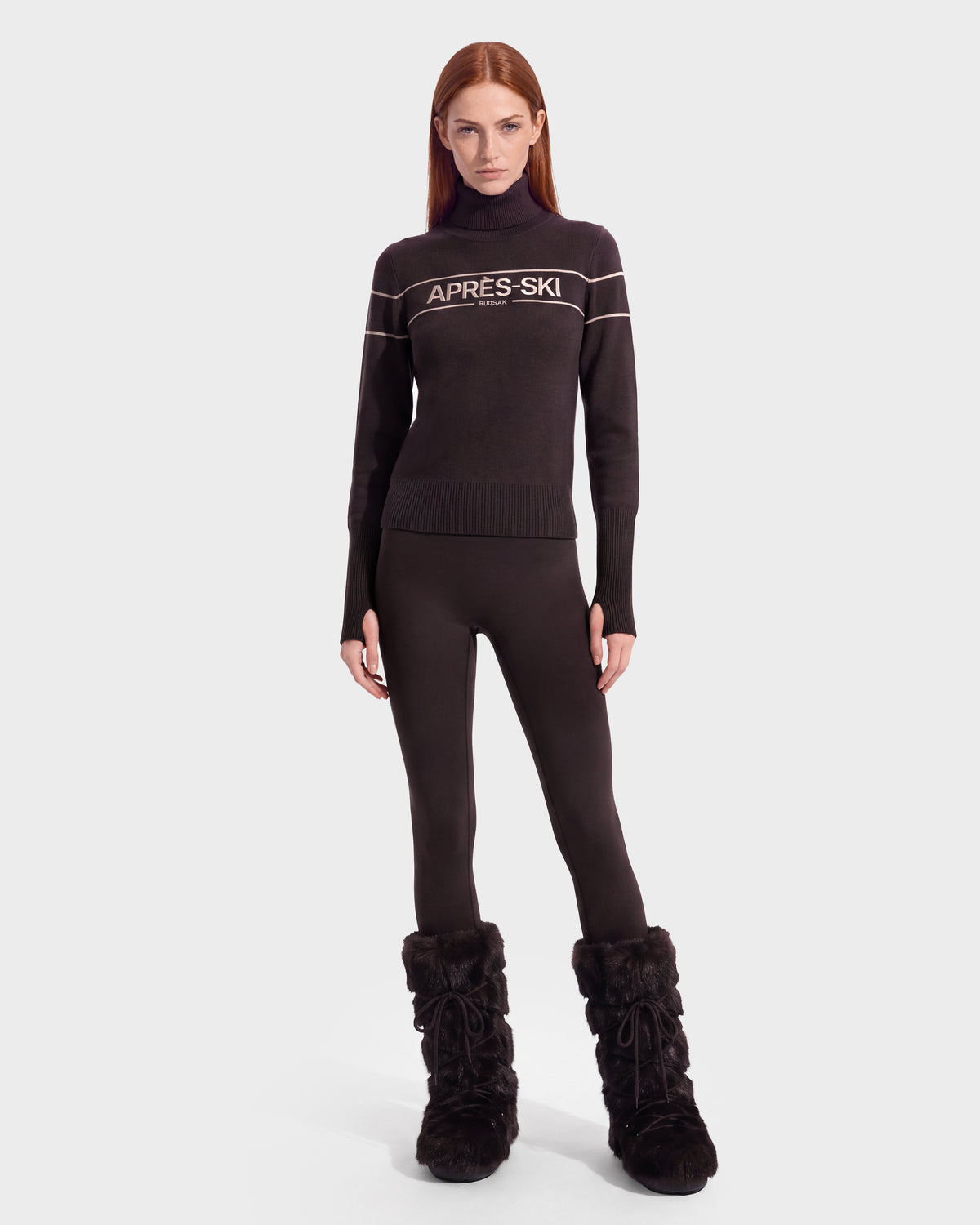 A woman with straight red hair showcases her winter wardrobe, wearing the Rudsak ERIKA - 8525705 Espresso turtleneck sweater, paired with black leggings and black furry boots, standing against a plain white background.