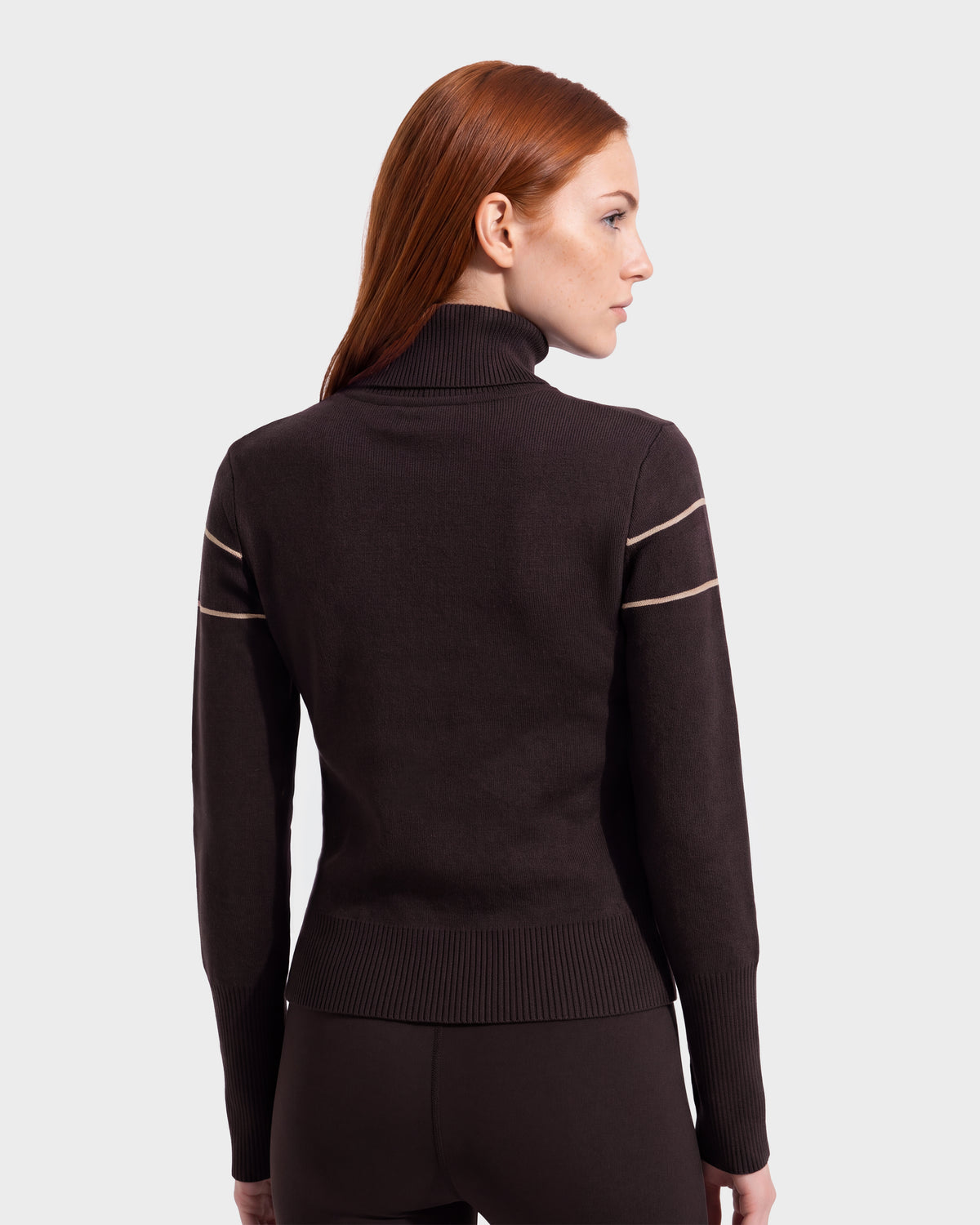 A woman with long red hair, standing and facing away against a plain light background, wears the Rudsak ERIKA - 8525705 Espresso après-ski turtleneck sweater featuring subtle stripes on the sleeves—an ideal piece for any winter wardrobe.