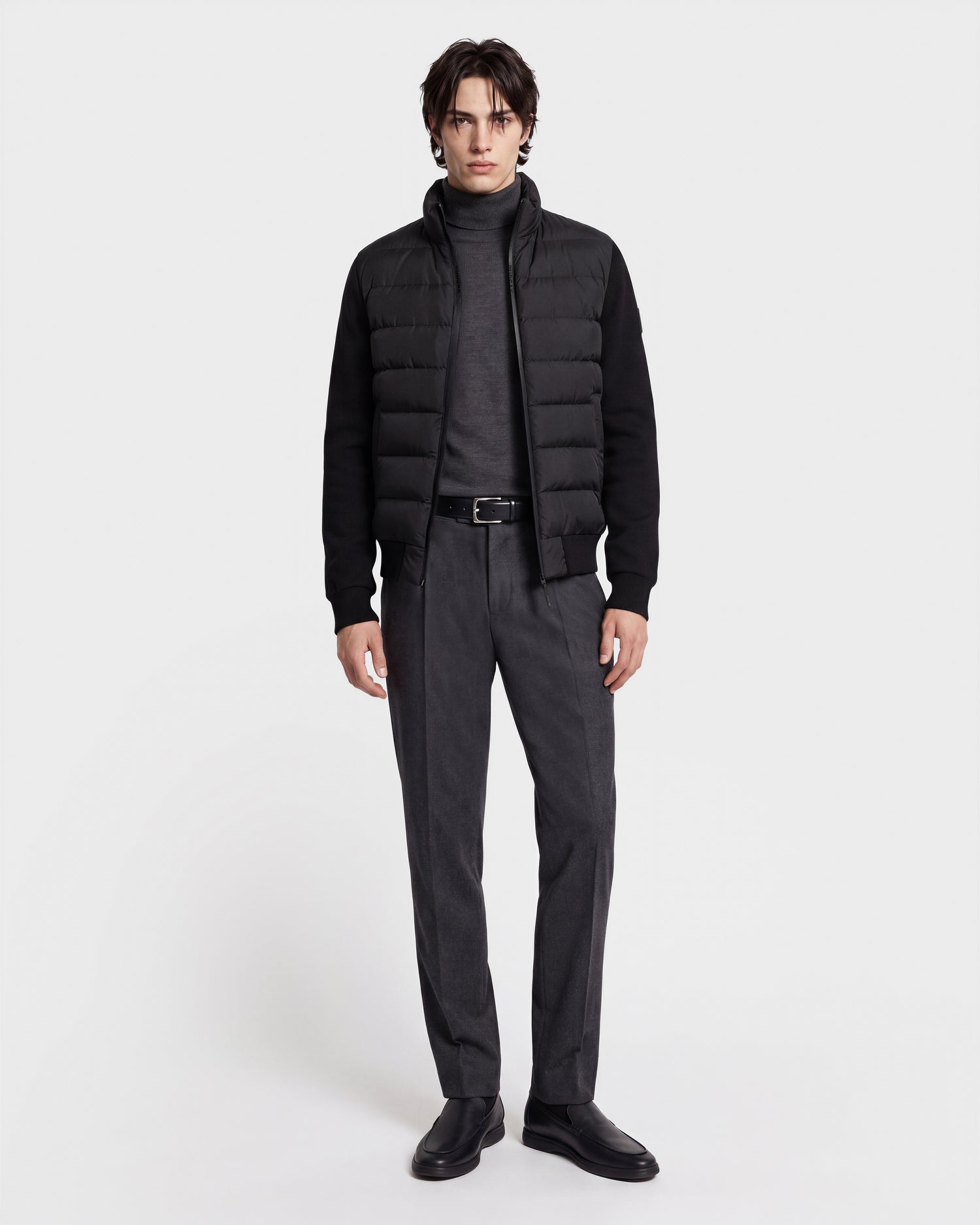 A person stands against a white background wearing the Rudsak EUGENE - 6124721 Black men's hybrid jacket crafted from performance luxury fabric, paired with a gray turtleneck, gray trousers, a belt, and black shoes.