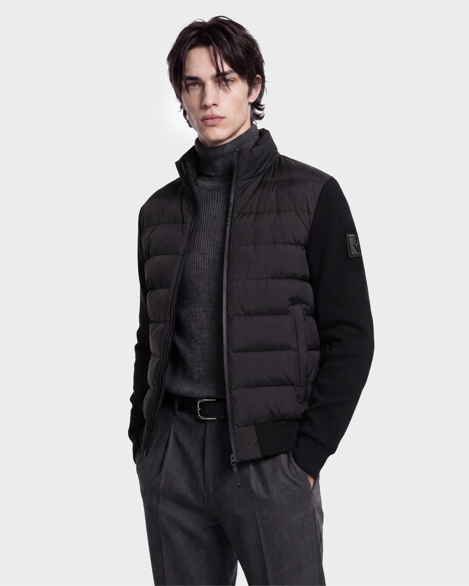 A man wearing the Rudsak EUGENE - 6124721 Black men's hybrid jacket, crafted from performance luxury fabric, over a gray turtleneck sweater and gray pants stands with his hands in his pockets against a plain light background.