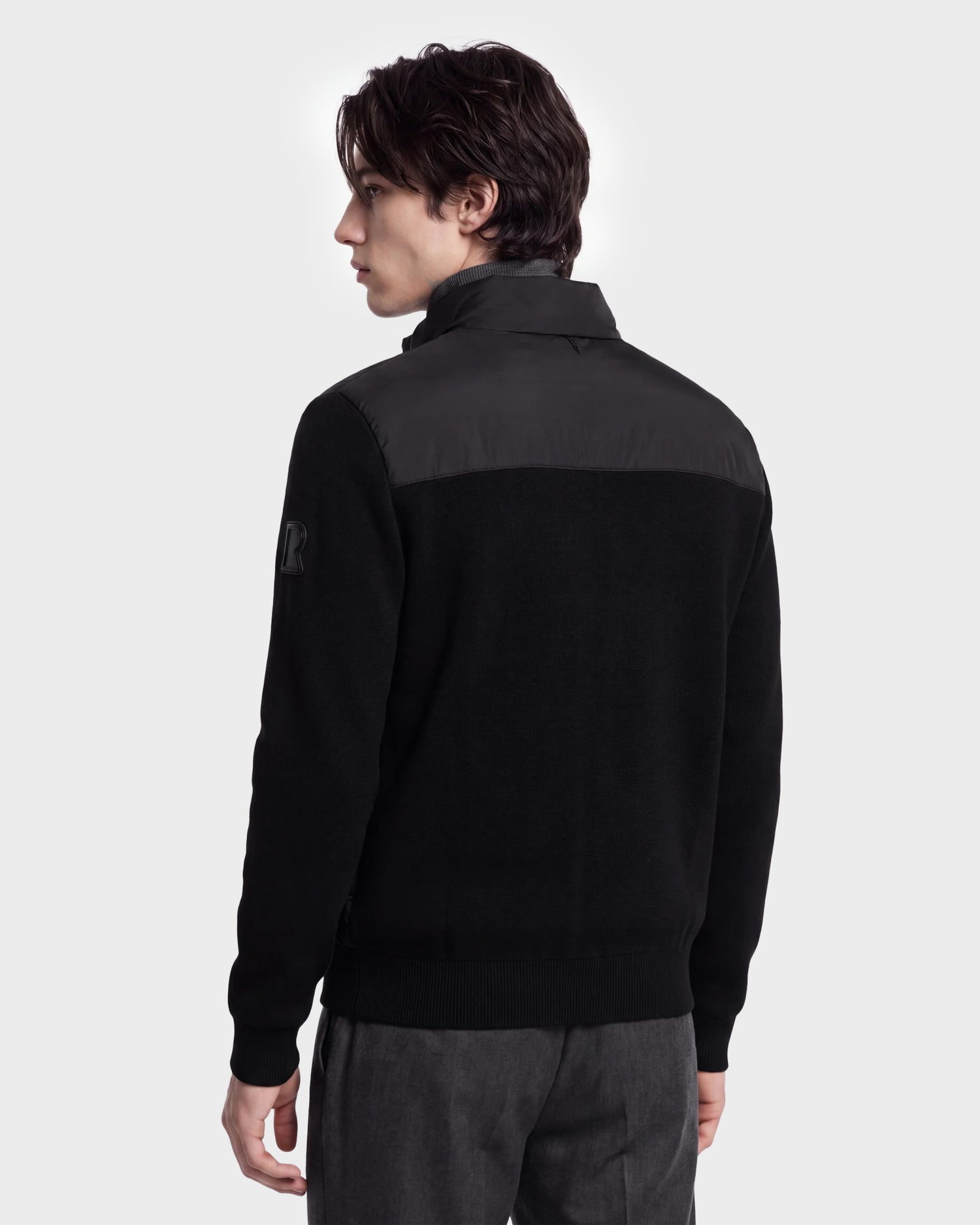 A person with dark hair is shown from the back, wearing the Rudsak EUGENE - 6124721 Black men’s hybrid jacket with a high collar and gray pants, standing against a plain light background.