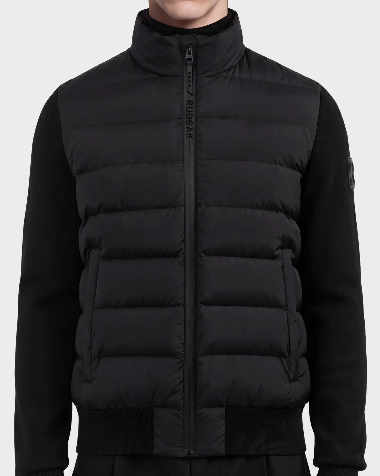 A person wearing the EUGENE - 6124721 Black men's hybrid jacket by Rudsak, featuring a high collar, zip closure with the Rudsak logo on the zipper flap, crafted from performance luxury fabric with ribbed cuffs and sides for a minimalist, modern look.