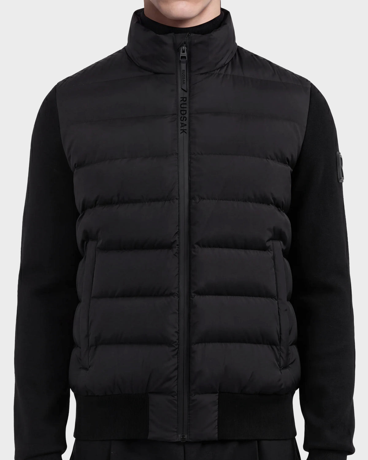 A person wearing the EUGENE - 6124721 Black men's hybrid jacket by Rudsak, featuring a high collar, zip closure with the Rudsak logo on the zipper flap, crafted from performance luxury fabric with ribbed cuffs and sides for a minimalist, modern look.