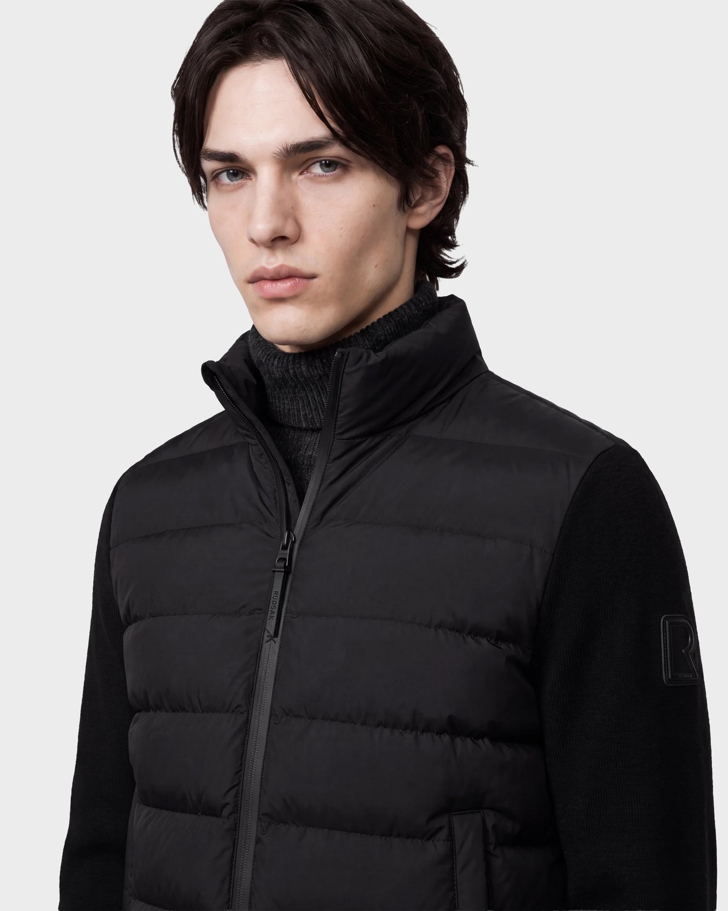 A young man with medium-length brown hair models the Rudsak EUGENE - 6124721 Black men's mixed-media jacket over a black turtleneck, gazing at the camera with a neutral expression against a plain light background.