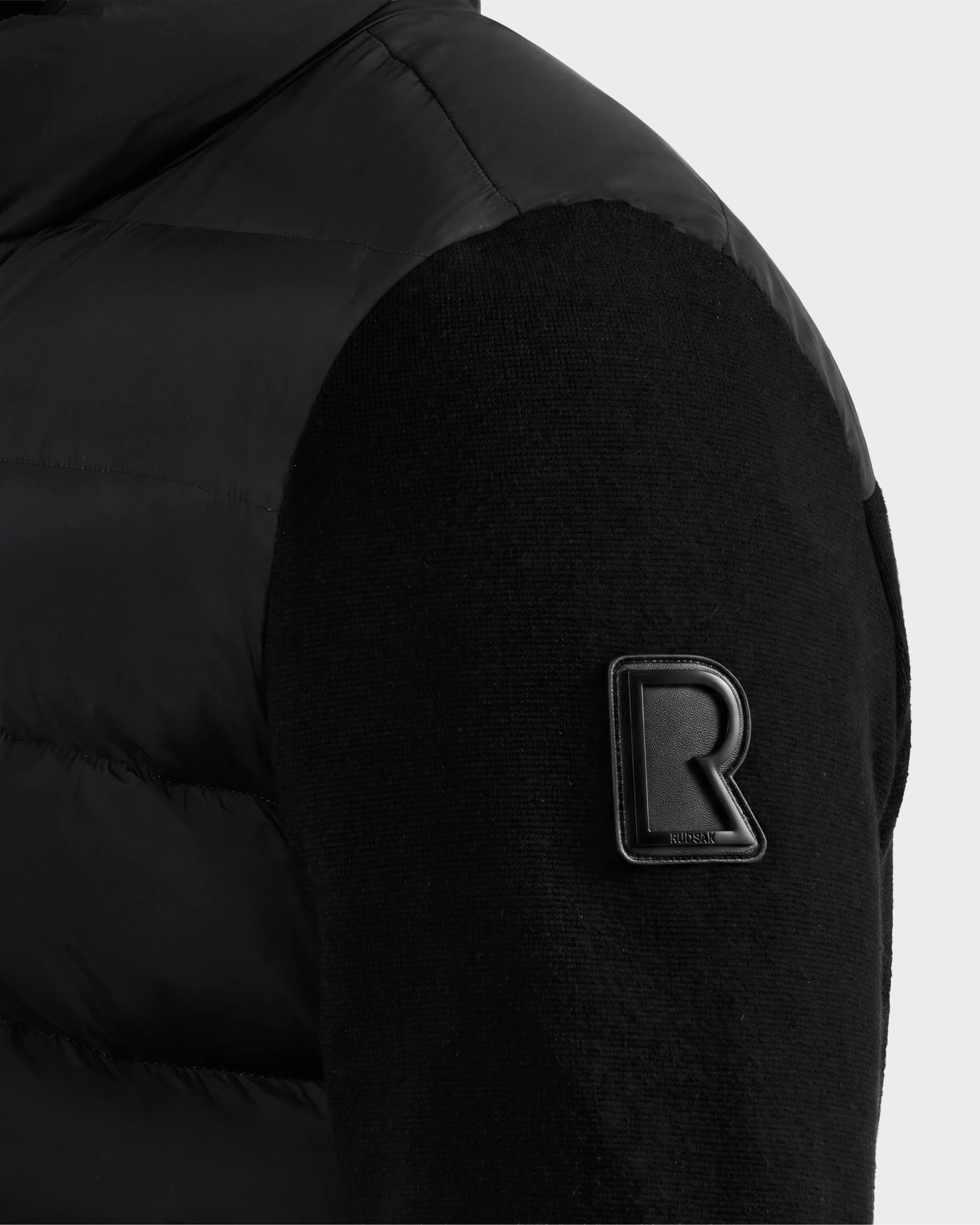 Close-up of the EUGENE - 6124721 Black jacket sleeve by Rudsak, showcasing a raised R logo patch near the shoulder, intricate stitched fabric pattern, and a sleek, shiny upper shoulder section.