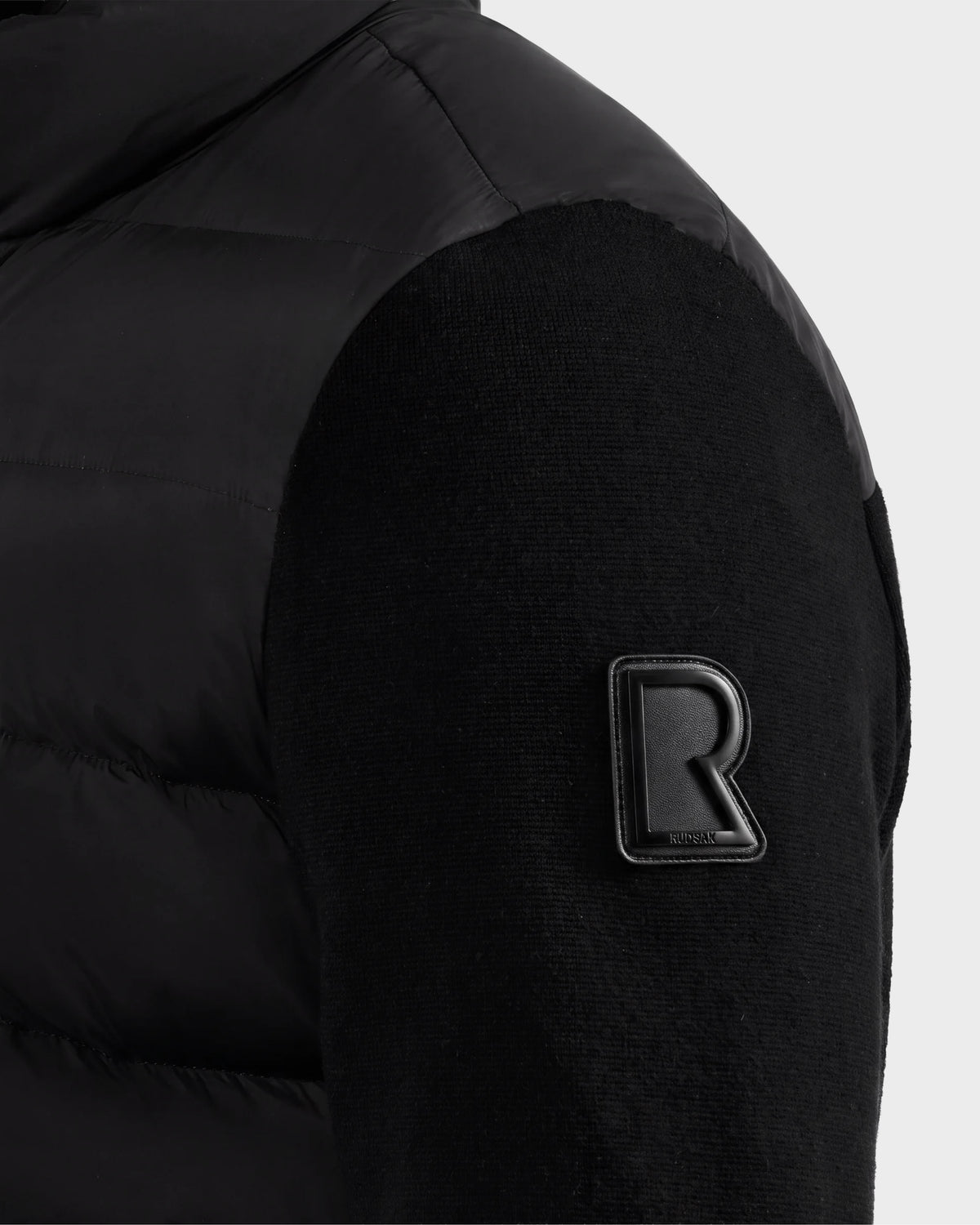 Close-up of the EUGENE - 6124721 Black jacket sleeve by Rudsak, showcasing a raised R logo patch near the shoulder, intricate stitched fabric pattern, and a sleek, shiny upper shoulder section.