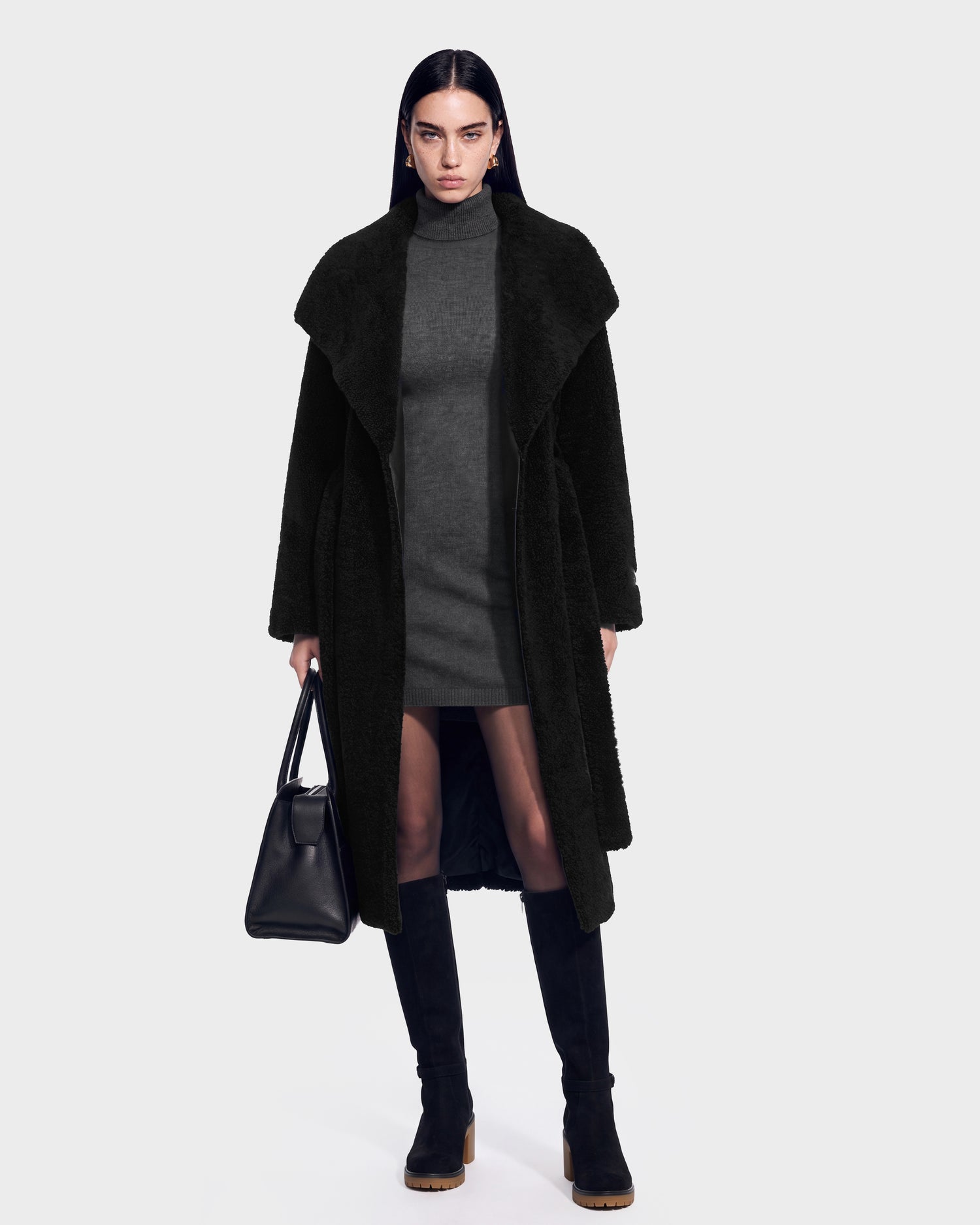 A woman with straight dark hair wears the Rudsak FRANCIE - 8126873 Black long coat over a gray dress, paired with black knee-high boots and holding a black handbag, standing against a plain light background. This item is available for pre-order and is expected to ship by September 15th.