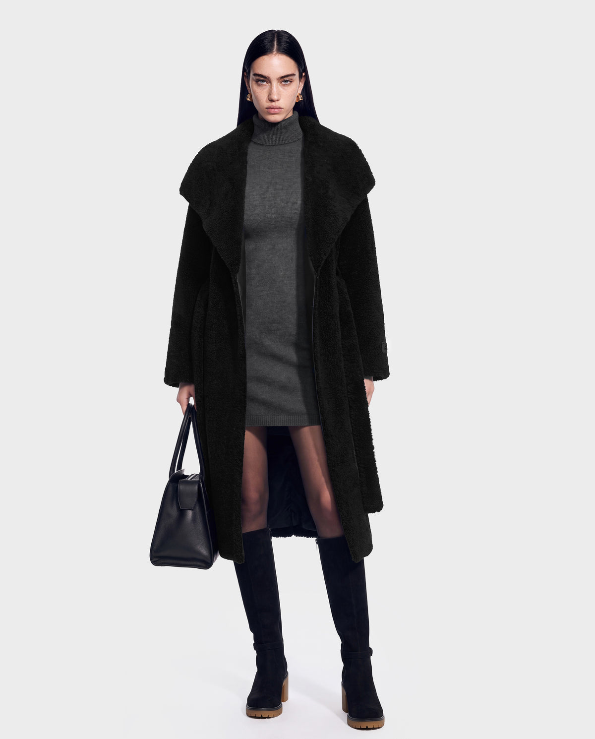 A woman stands against a plain light background, wearing the Rudsak FRANCIE - 8126873 long black vegan leather coat over a gray dress, paired with knee-high black boots and holding a black handbag. She has straight dark hair and a serious expression.