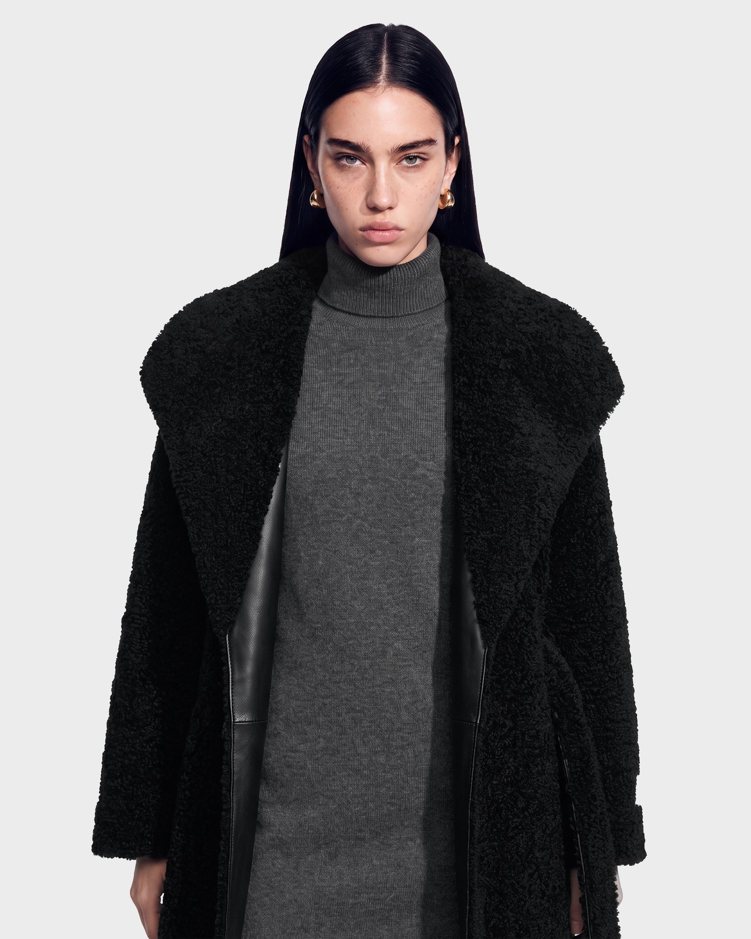 A woman with straight dark hair models the FRANCIE - 8126873 Black textured coat by Rudsak over a gray turtleneck dress, standing against a plain light background and looking directly at the camera with a neutral expression. This item is available for pre-order and is expected to ship by September 15th.