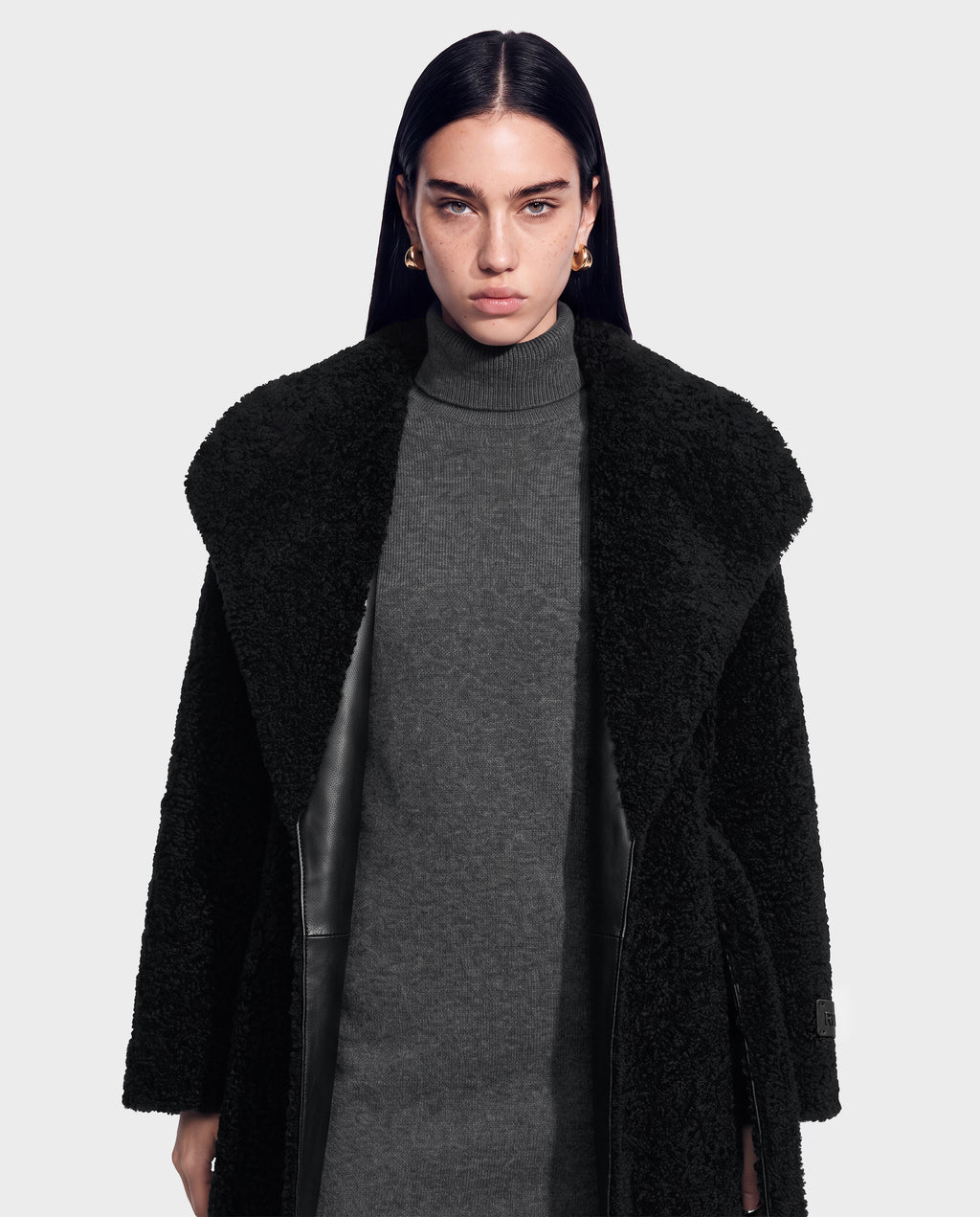 A woman with straight dark hair, wearing gold hoop earrings, a grey turtleneck dress, and the FRANCIE - 8126873 Black textured vegan leather coat by Rudsak, stands against a plain light background, looking directly at the camera with a neutral expression.