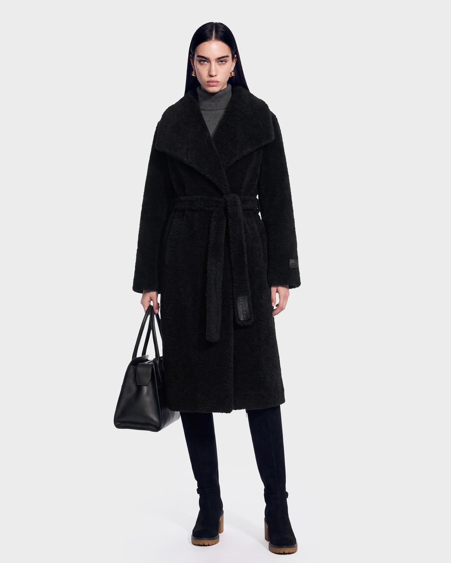 A woman wearing the FRANCIE - 8126873 Black long belted coat by Rudsak, paired with a gray turtleneck and black boots, holds a black handbag. She stands facing forward against a plain white background. This stylish look is available for pre-order and is expected to ship by September 15th.