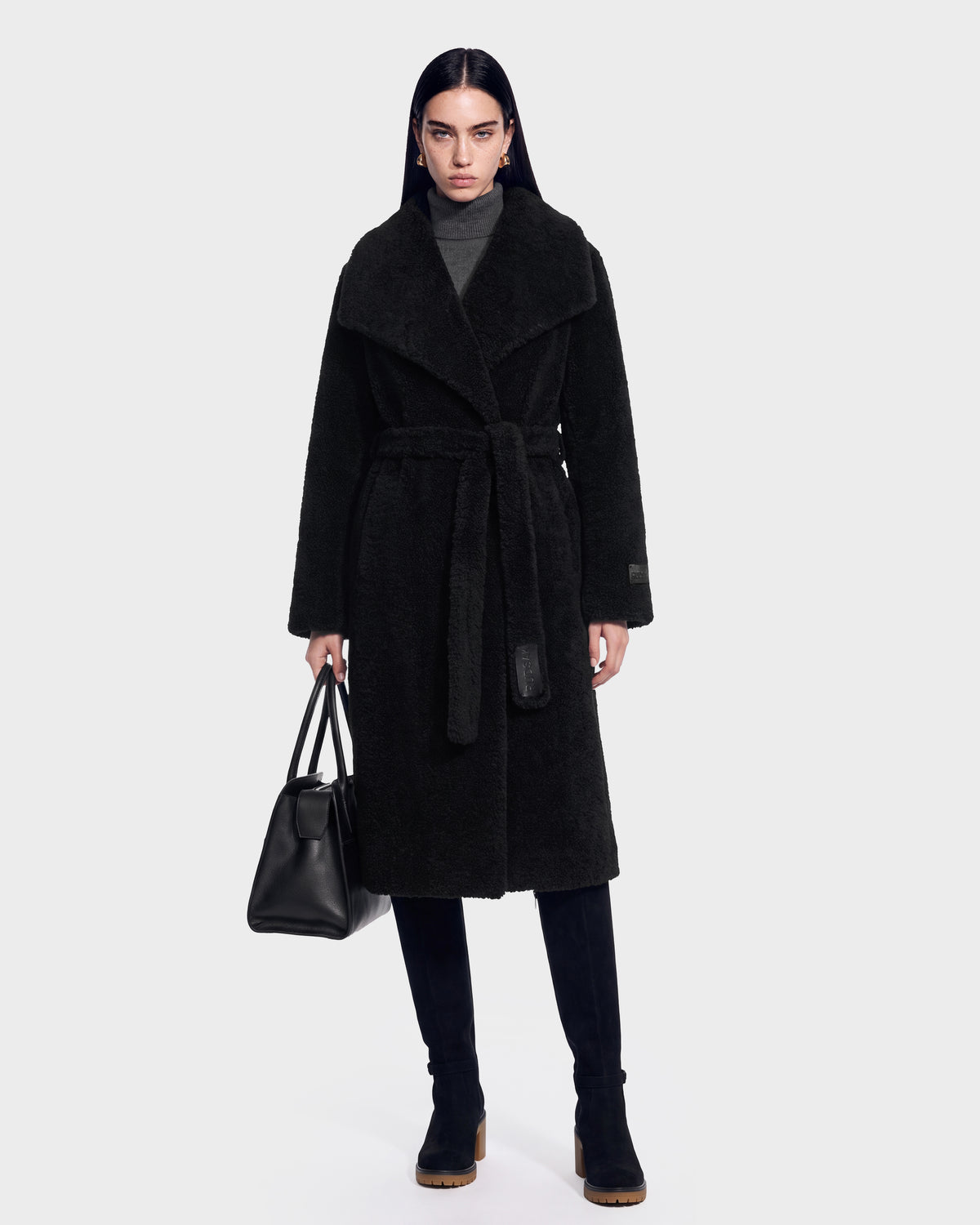 A woman wearing the FRANCIE - 8126873 Black long belted coat by Rudsak, paired with a gray turtleneck and black boots, holds a black handbag. She stands facing forward against a plain white background. This stylish look is available for pre-order and is expected to ship by September 15th.