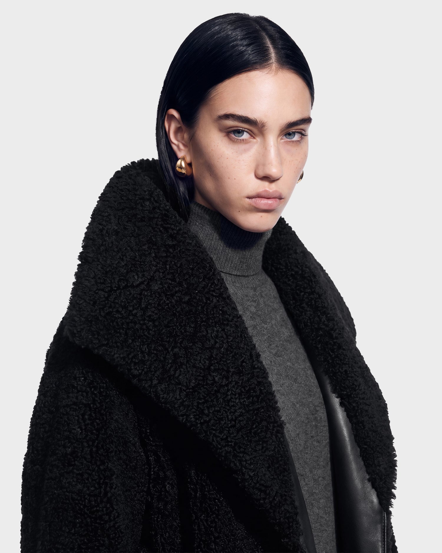 A person with straight dark hair, gold hoop earrings, and piercing blue eyes wears the FRANCIE - 8126873 Black textured coat by Rudsak, featuring a large collar over a gray turtleneck, posing against a plain light background—an effortlessly modern look now available for pre-order and expected to ship by September 15th.