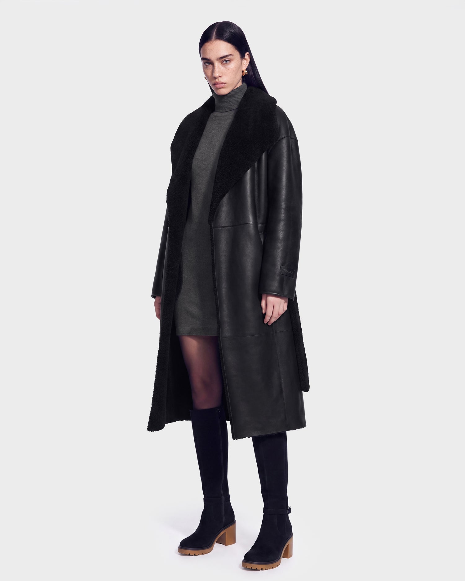 A woman stands against a plain light background wearing the FRANCIE - 8126873 Black shearling coat by Rudsak, styled with a gray turtleneck dress, black tights, and black heeled boots. This chic look is available for pre-order and is expected to ship by September 15th.