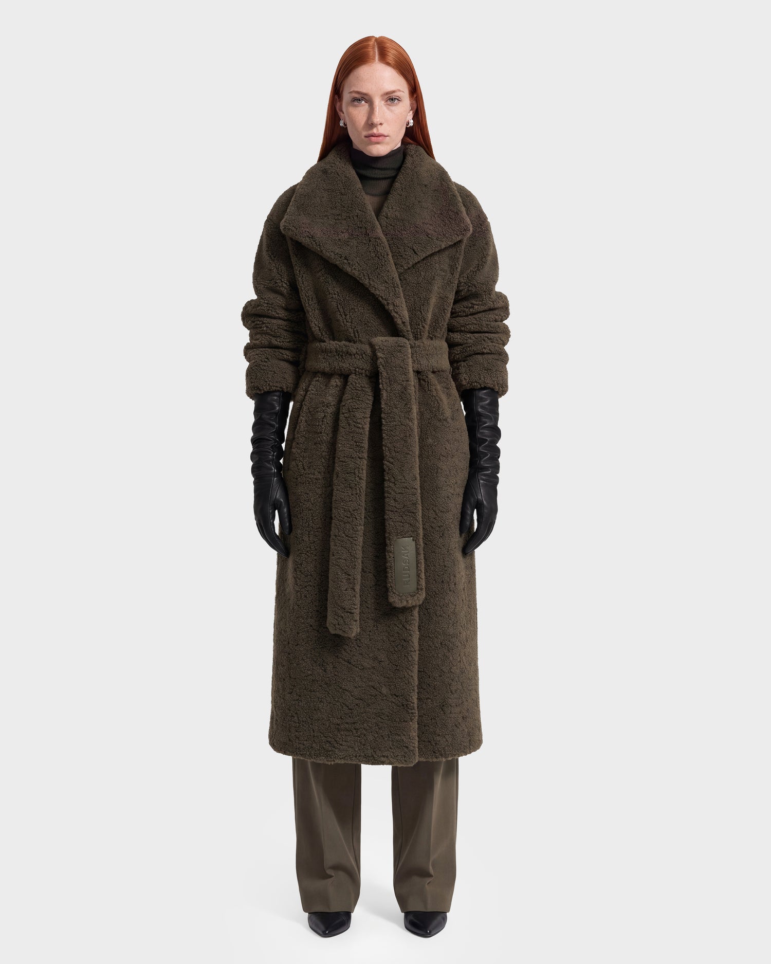 A woman with long red hair models the Rudsak FRANCIE - 8126873 Olive, a long, textured dark brown belted coat, layered over dark pants with black gloves and shoes. Photographed against a plain white background, this piece is available now for pre-order and expected to ship by September 15th.