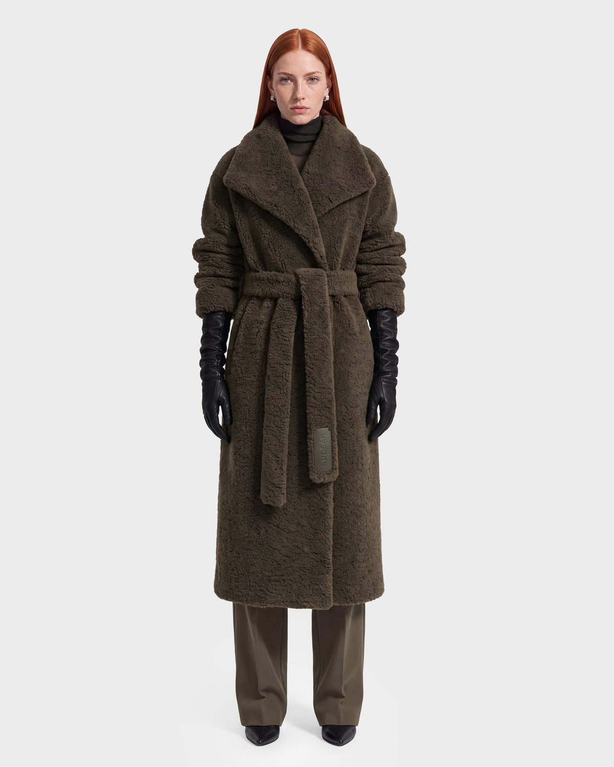 A woman with long red hair models the Rudsak FRANCIE - 8126873 Olive, a long, textured dark brown belted coat, layered over dark pants with black gloves and shoes. Photographed against a plain white background, this piece is available now for pre-order and expected to ship by September 15th.