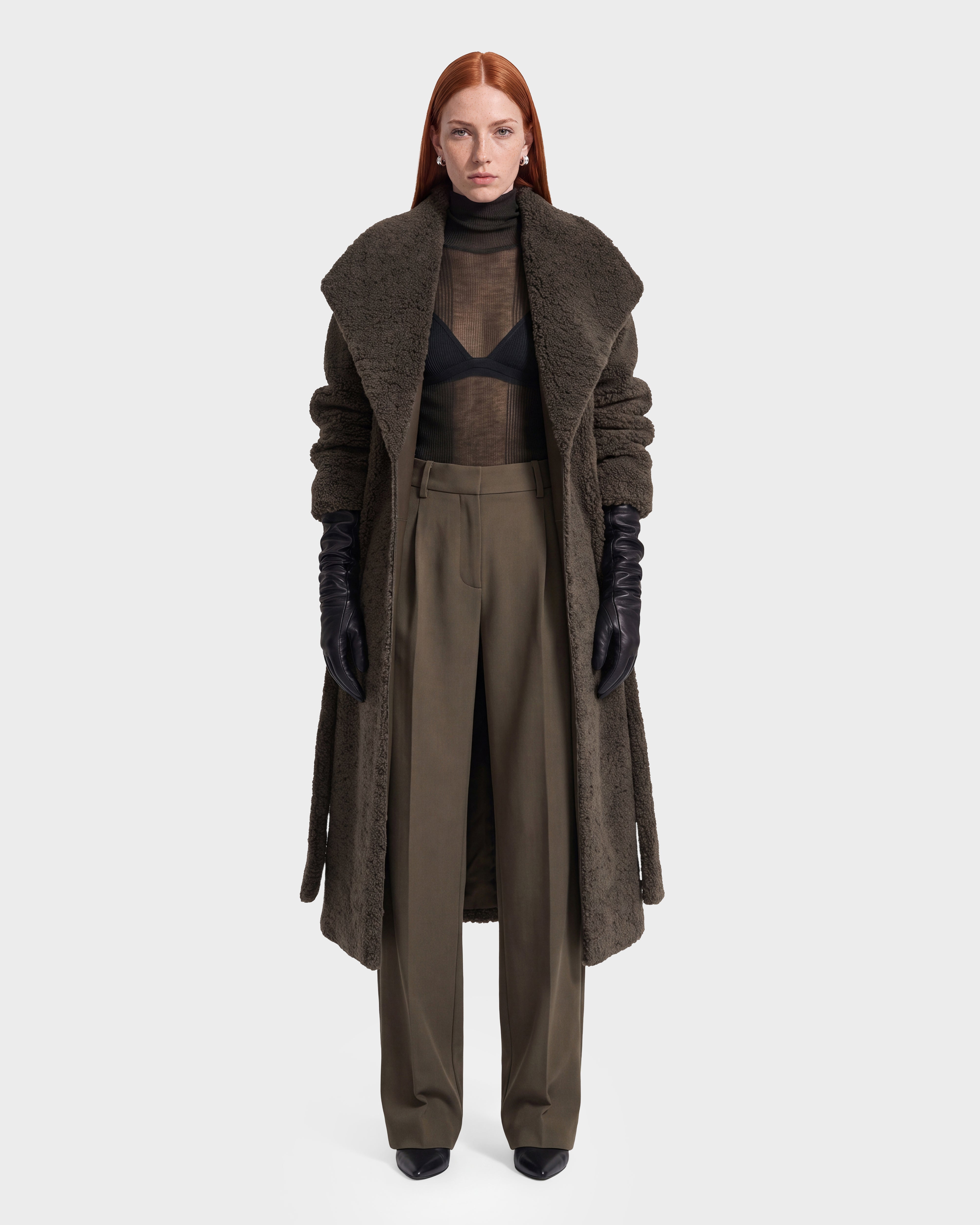 A person with long red hair wears a long brown textured coat, black gloves, a sheer black top with a visible bra, and the Rudsak FRANCIE - 8126873 Olive high-waisted wide-leg pants, standing against a plain white background. This striking look is available for pre-order now and is expected to ship by September 15th.