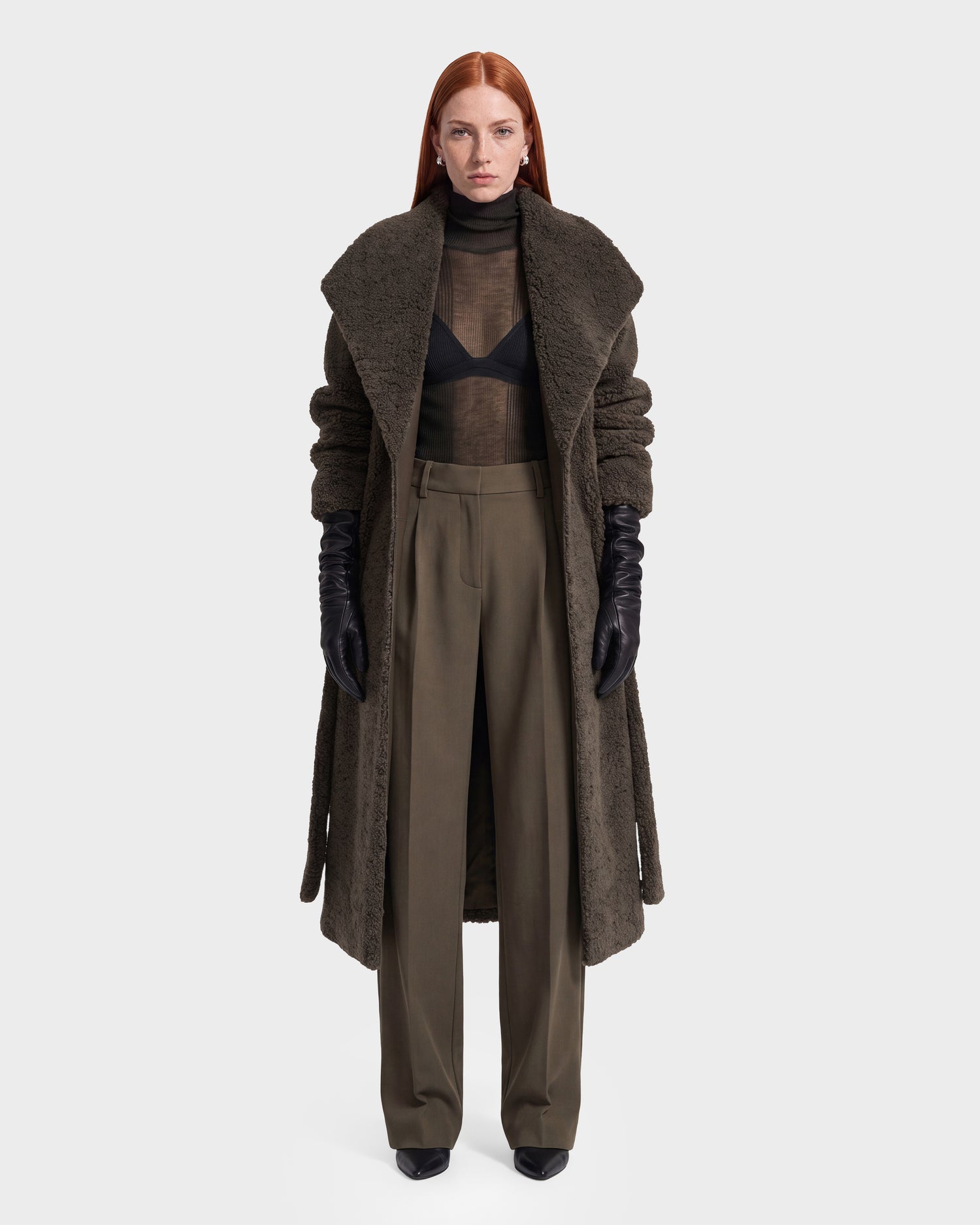 A person with long red hair wears a long brown textured coat, black gloves, a sheer black top with a visible bra, and the Rudsak FRANCIE - 8126873 Olive high-waisted wide-leg pants, standing against a plain white background. This striking look is available for pre-order now and is expected to ship by September 15th.