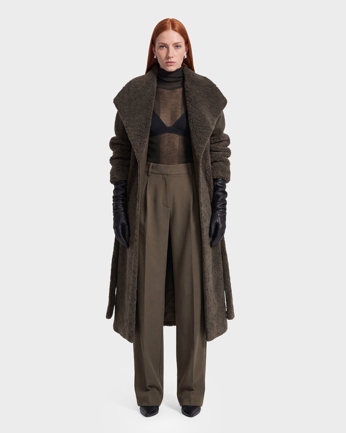 A person with long red hair wears a long brown textured coat, black gloves, a sheer black top with a visible bra, and the Rudsak FRANCIE - 8126873 Olive high-waisted wide-leg pants, standing against a plain white background. This striking look is available for pre-order now and is expected to ship by September 15th.