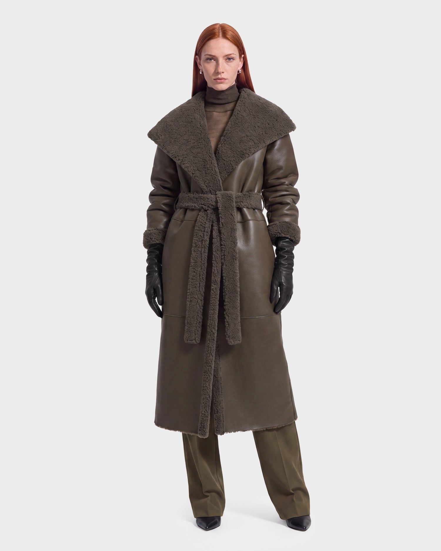 A woman with long red hair models the FRANCIE - 8126873 Olive shearling coat by Rudsak, featuring a wide collar, belted waist, matching pants, black gloves, and black pointed shoes, standing against a plain white background. This stylish ensemble is available for pre-order now and is expected to ship by September 15th.
