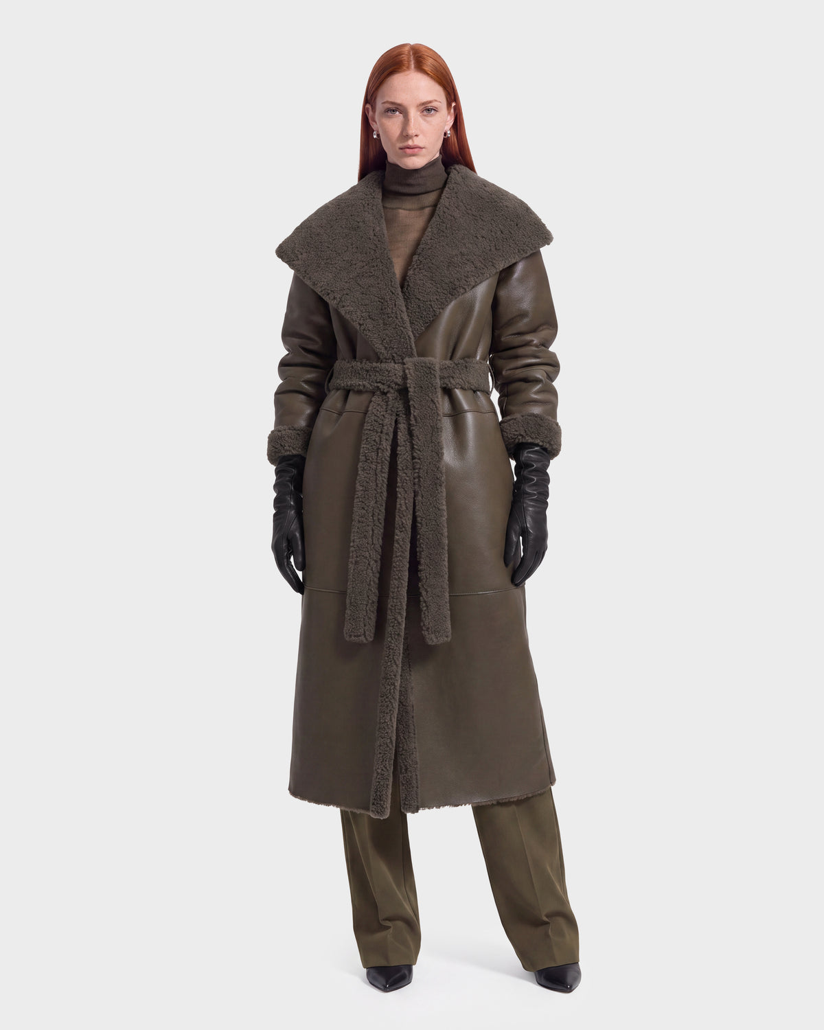 A woman with long red hair models the FRANCIE - 8126873 Olive shearling coat by Rudsak, featuring a wide collar, belted waist, matching pants, black gloves, and black pointed shoes, standing against a plain white background. This stylish ensemble is available for pre-order now and is expected to ship by September 15th.